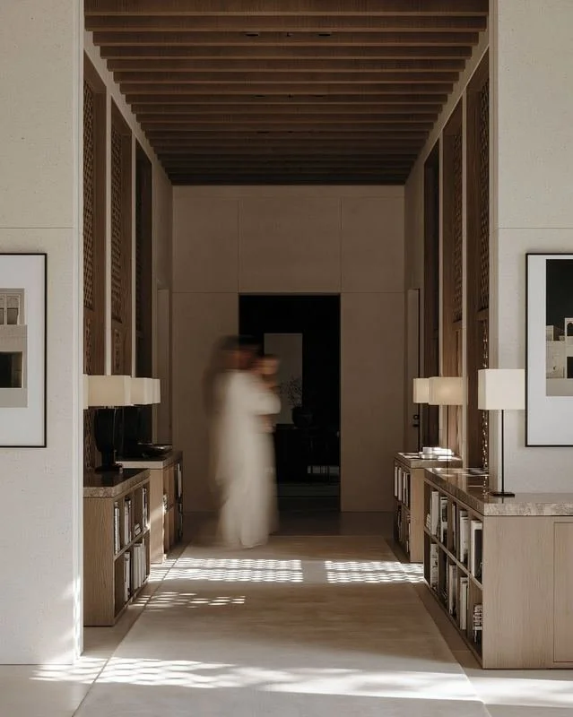 M18 · Árdh, concept image — a long timber-ceilinged corridor lined with low shelves and lamps, sunlight patterned on the floor, a figure in white walking through