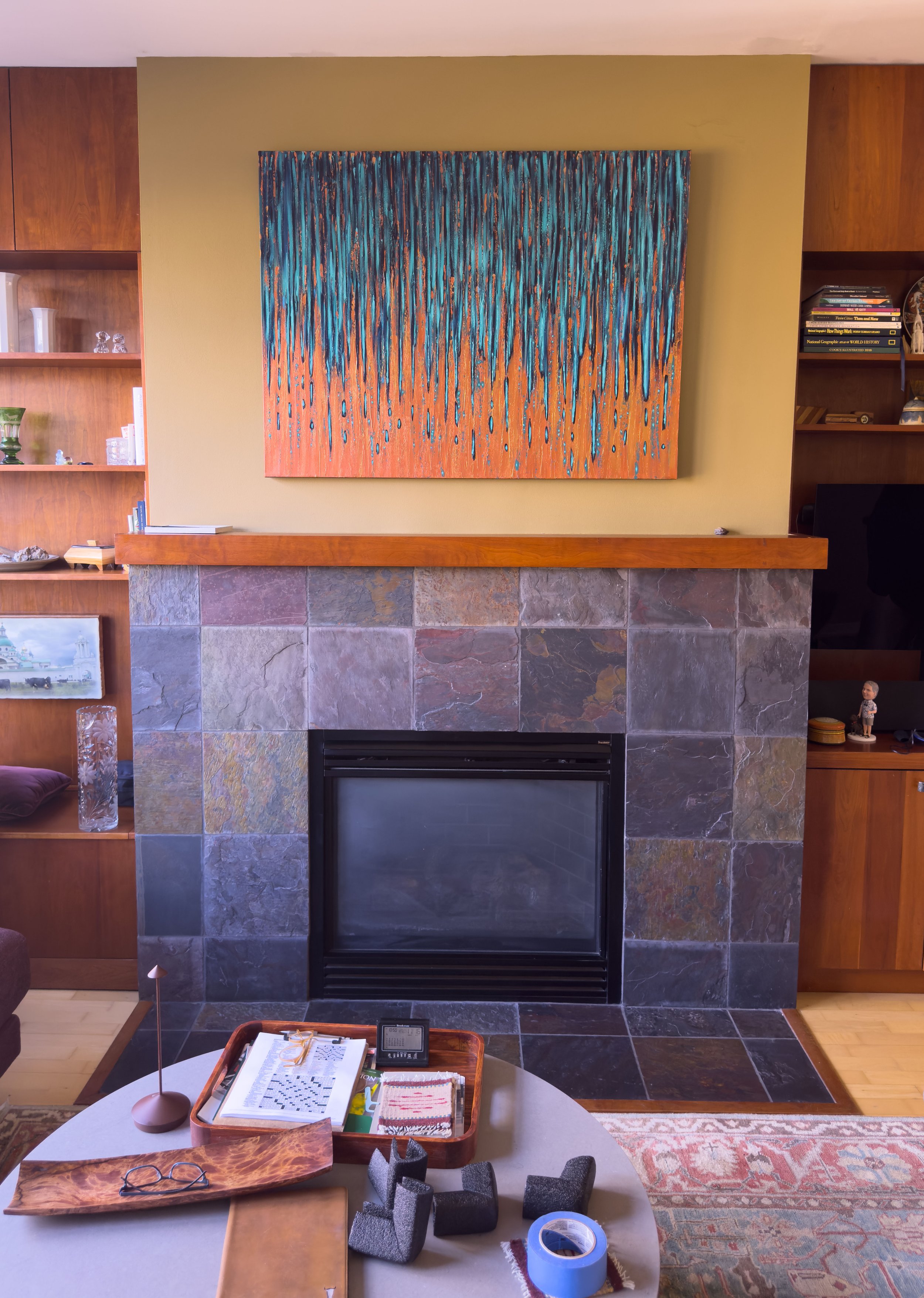 Living room with a fireplace, colorful abstract painting above it, and wooden bookshelves on either side, with a coffee table in front holding books, glasses, and office supplies.
