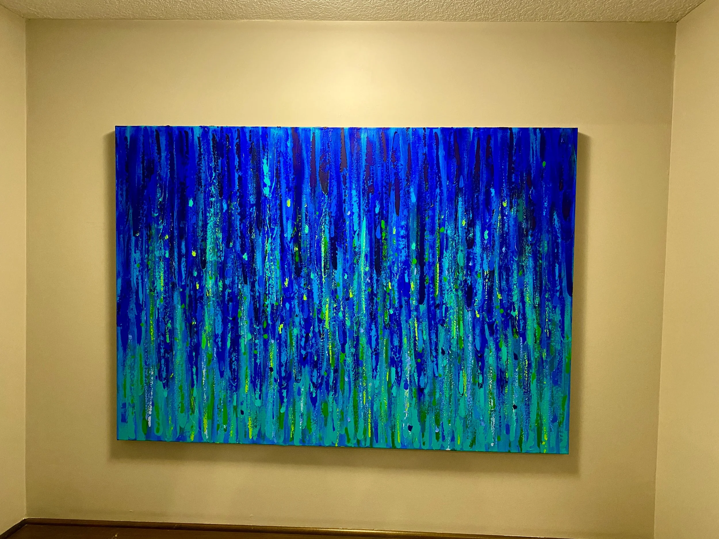 Abstract painting with vertical blue, green, and yellow streaks on a large canvas, hanging on a beige wall