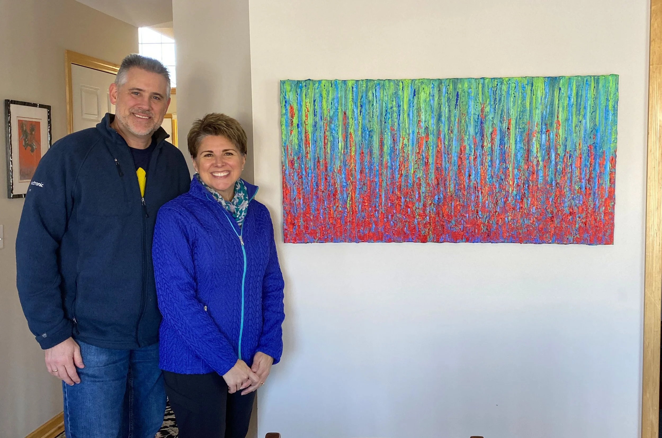 A smiling man and woman stand inside a house next to a colorful, abstract painting on the wall.