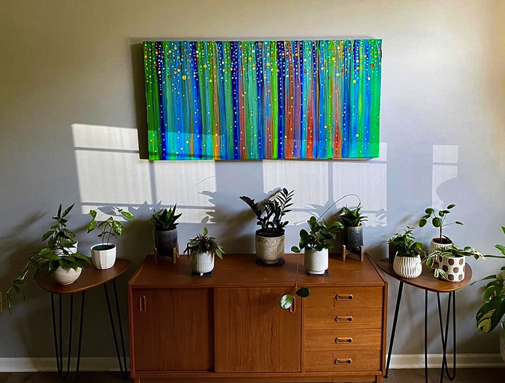 A wooden sideboard with five potted plants on top, flanked by two small wooden tables with additional plants, and a colorful abstract painting on the wall with shadows of window frames.