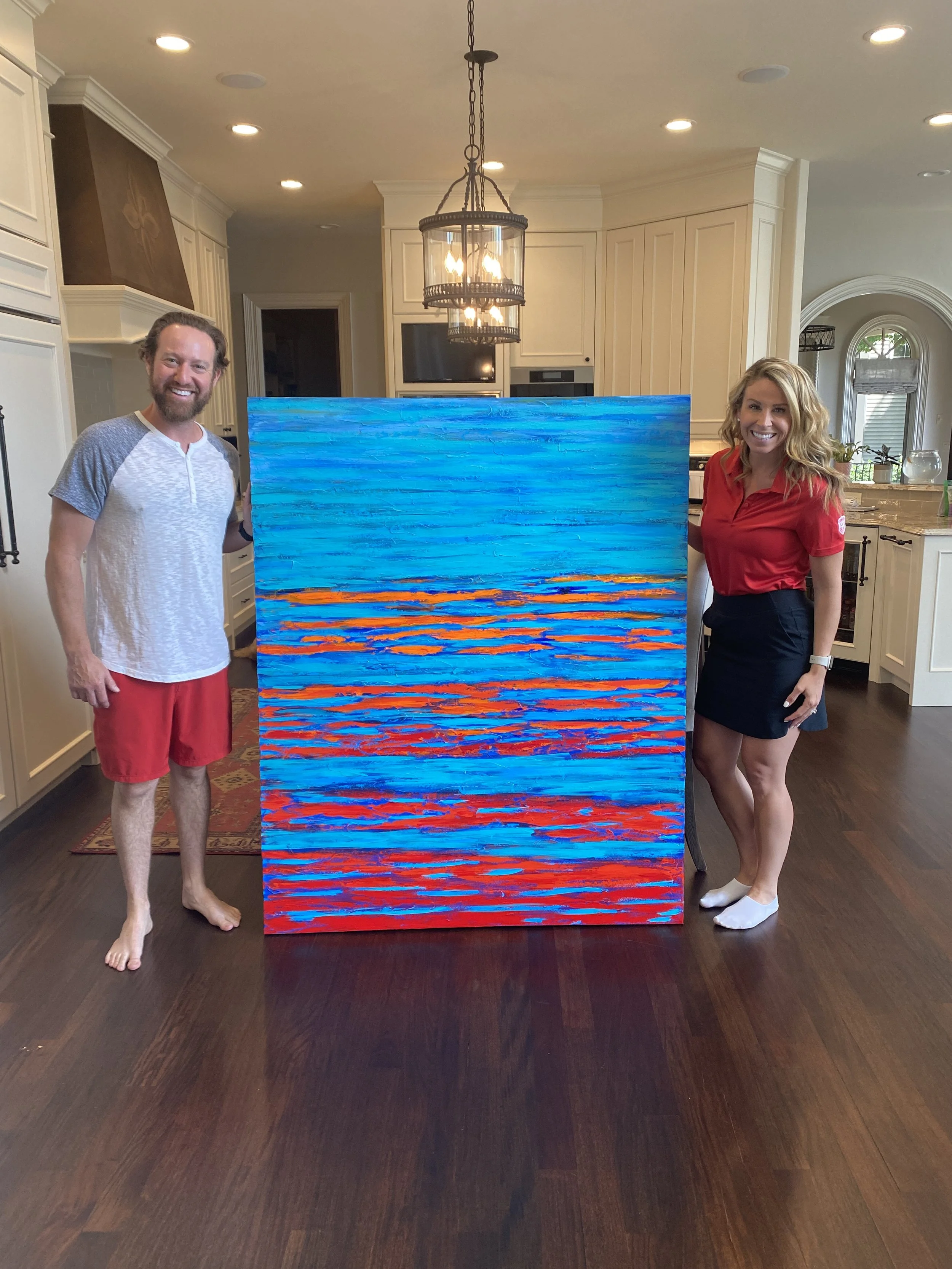 A man and a woman standing by a large abstract painting of a colorful sunset scene, inside a home kitchen with dark hardwood floors and white cabinetry.
