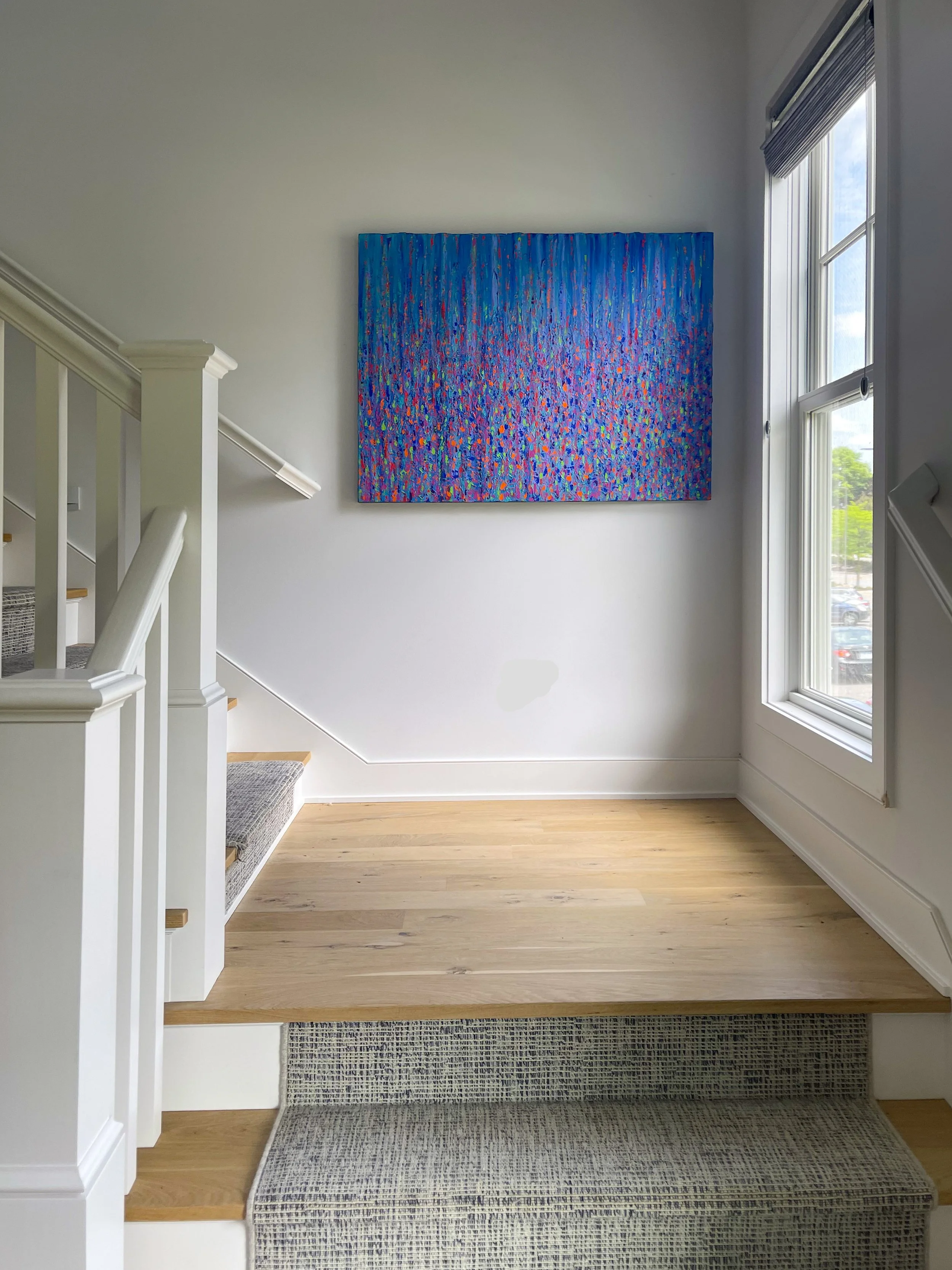 An interior view of a bright room with a staircase, a large colorful abstract painting on the wall, a window with blinds, and a wooden floor.
