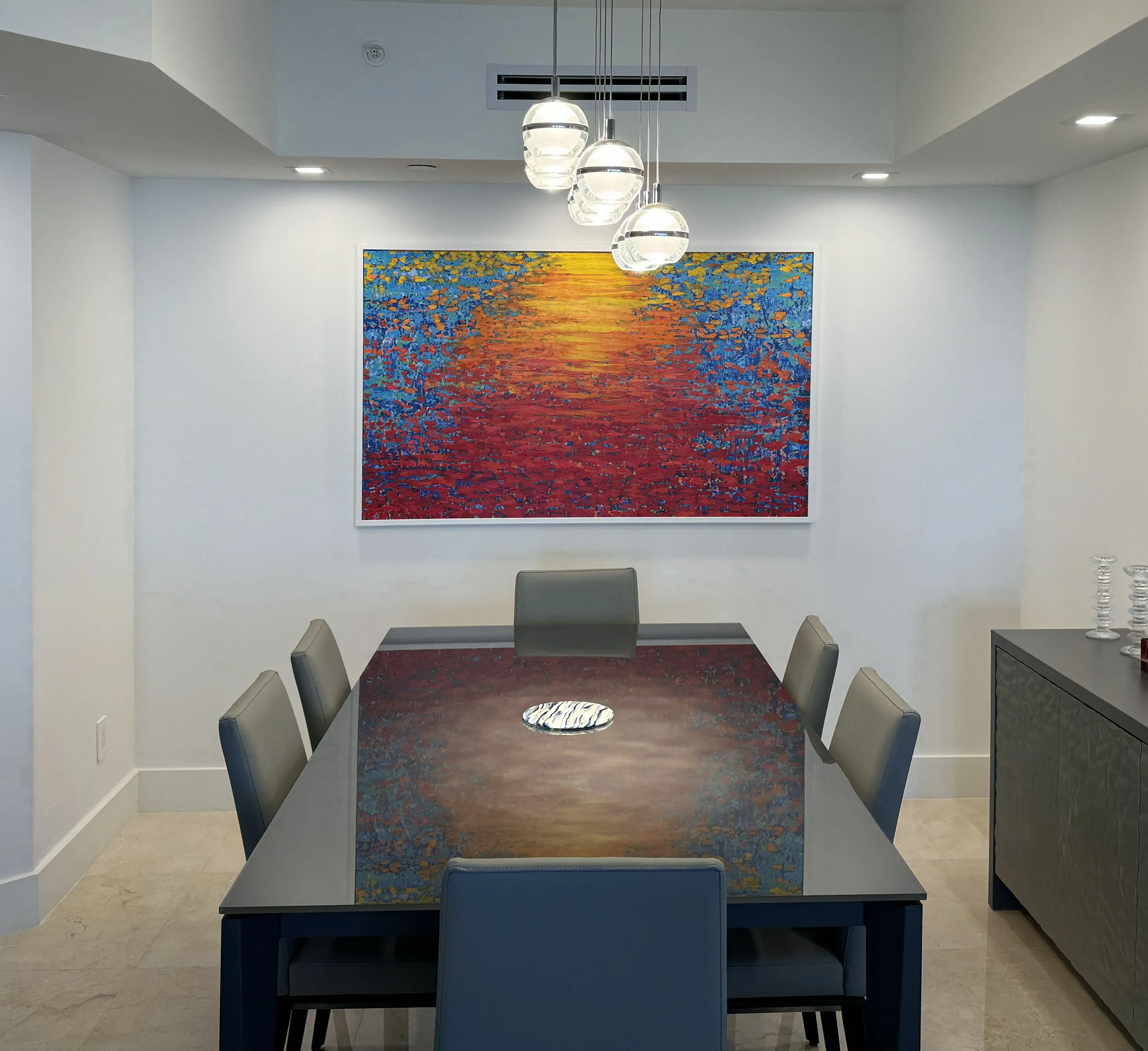 Modern dining room with a black table, six gray chairs, a colorful abstract painting of a sunset over water hanging on a white wall, and a sideboard with glassware. Lighting includes hanging pendant lights and recessed ceiling lights.