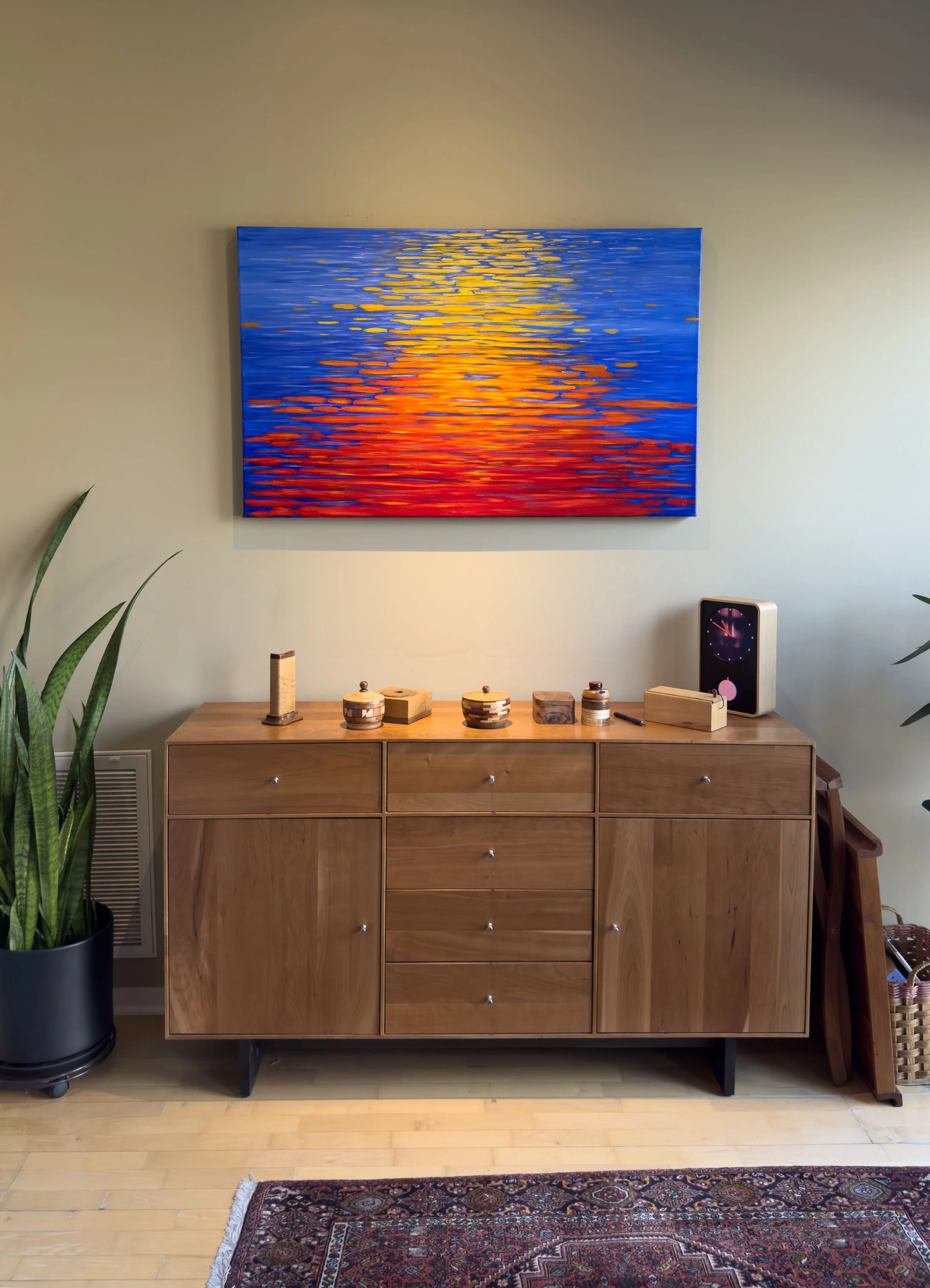 A wooden sideboard with decorative objects on top, a large colorful sunset landscape painting above, and a digital clock on the right, with potted plants on both sides and a patterned rug on the floor.
