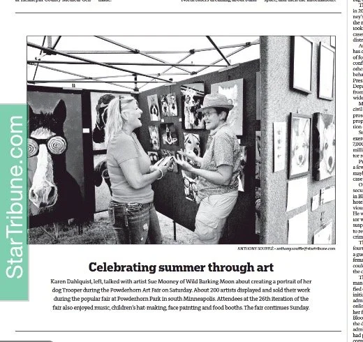Two women interacting at an outdoor art fair, with artwork displayed on boards behind them.