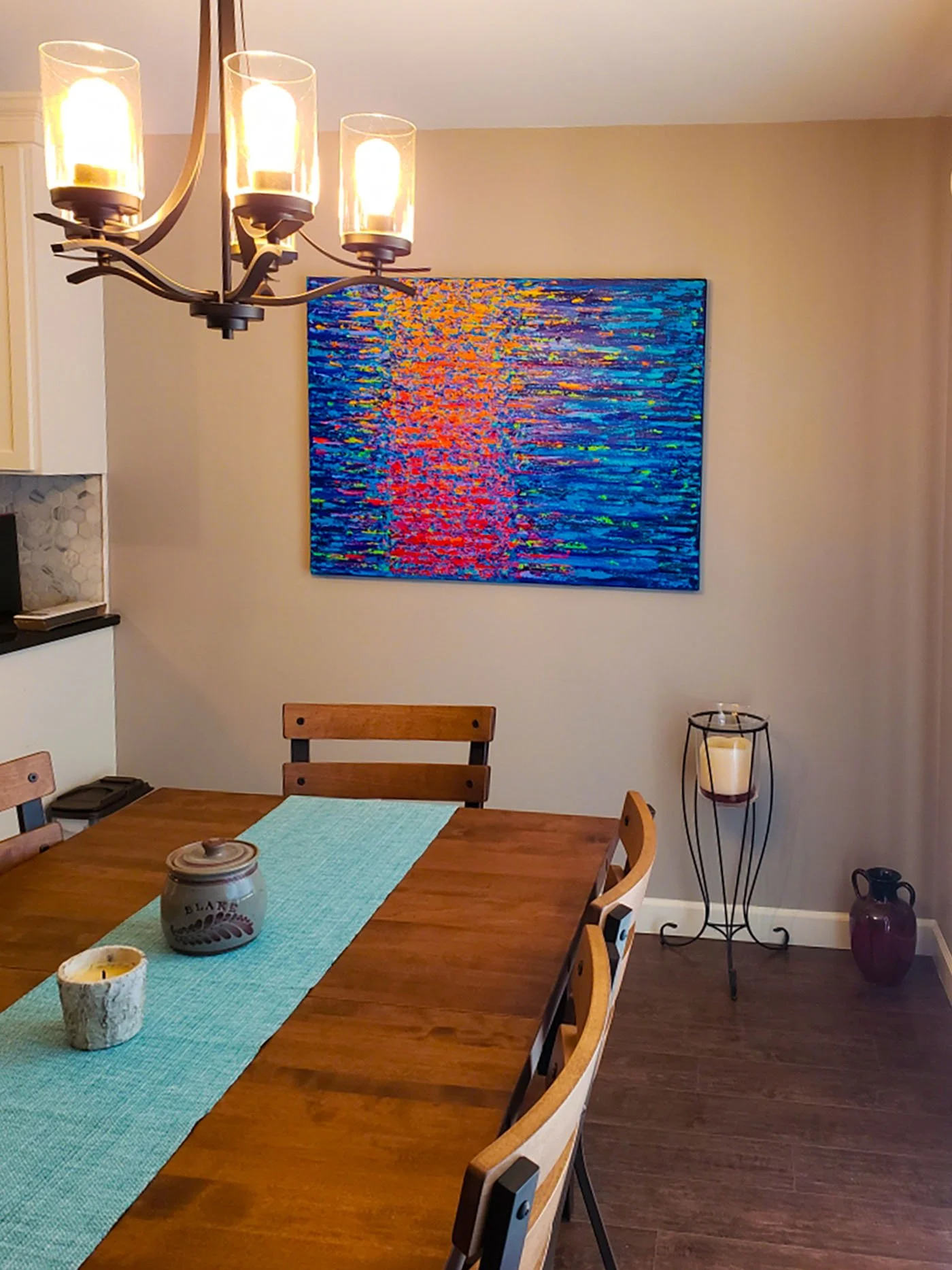 A dining room with a wooden table, four chairs, a blue table runner, candles, a large colorful abstract painting on the wall, and a floor candle holder.
