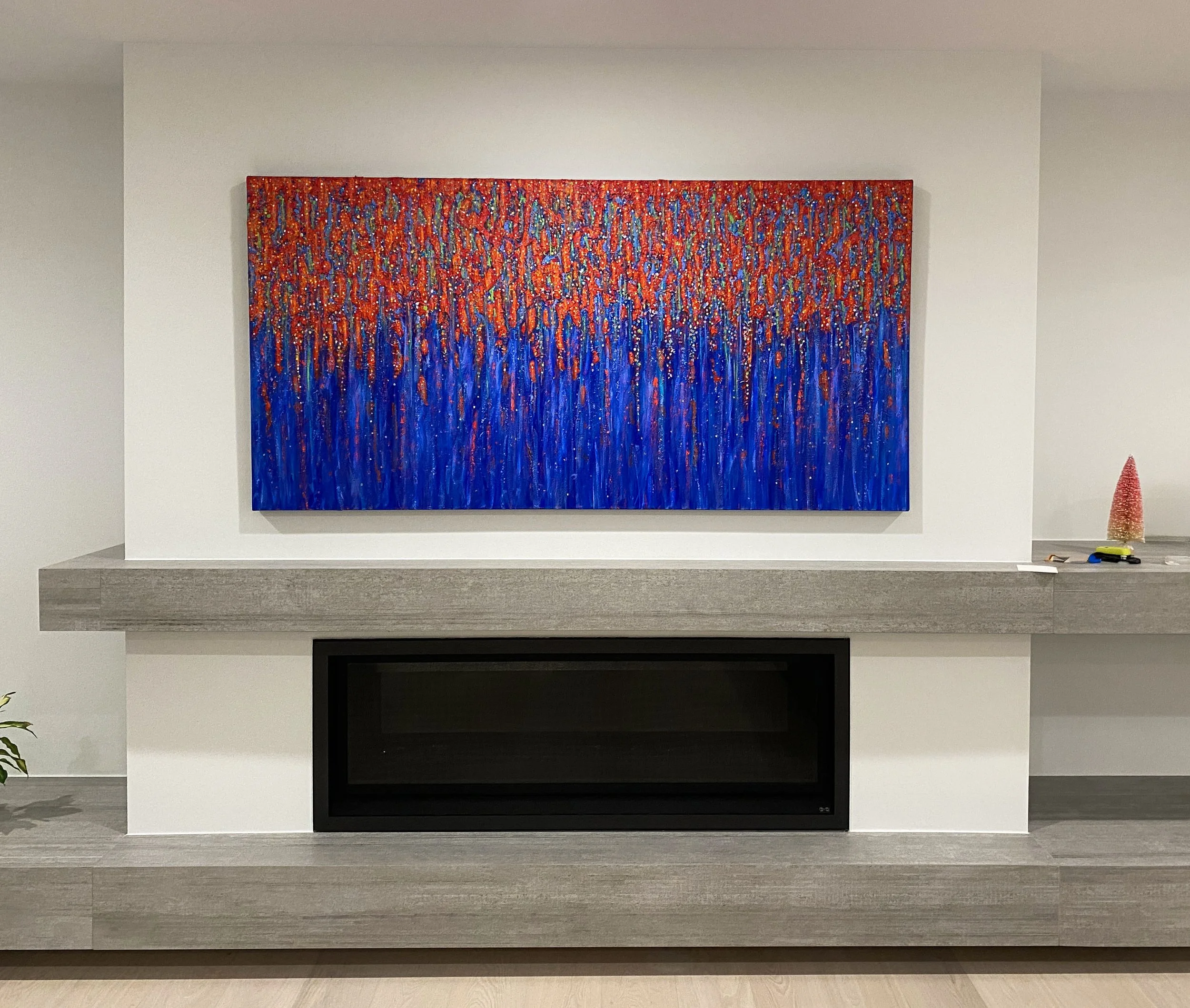 Abstract painting with red, orange, and yellow dots on top and vertical blue streaks below, hanging above a modern fireplace with a gray mantel in a white wall living room.