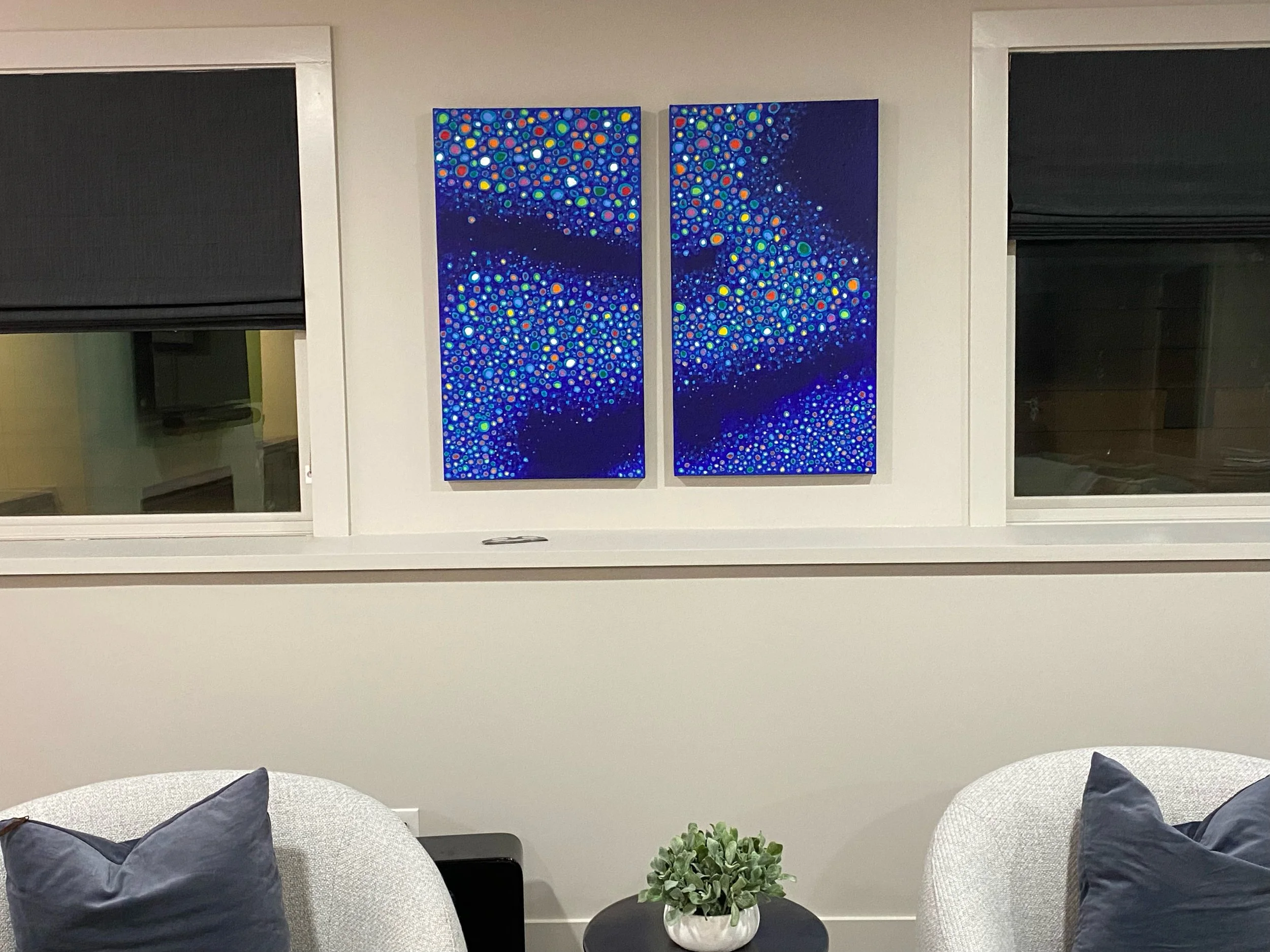 Two abstract paintings with blue, green, orange, and white dots in vertical panels on a wall above a white couch with dark pillows, a small potted plant on a black table, and windows with black shades.