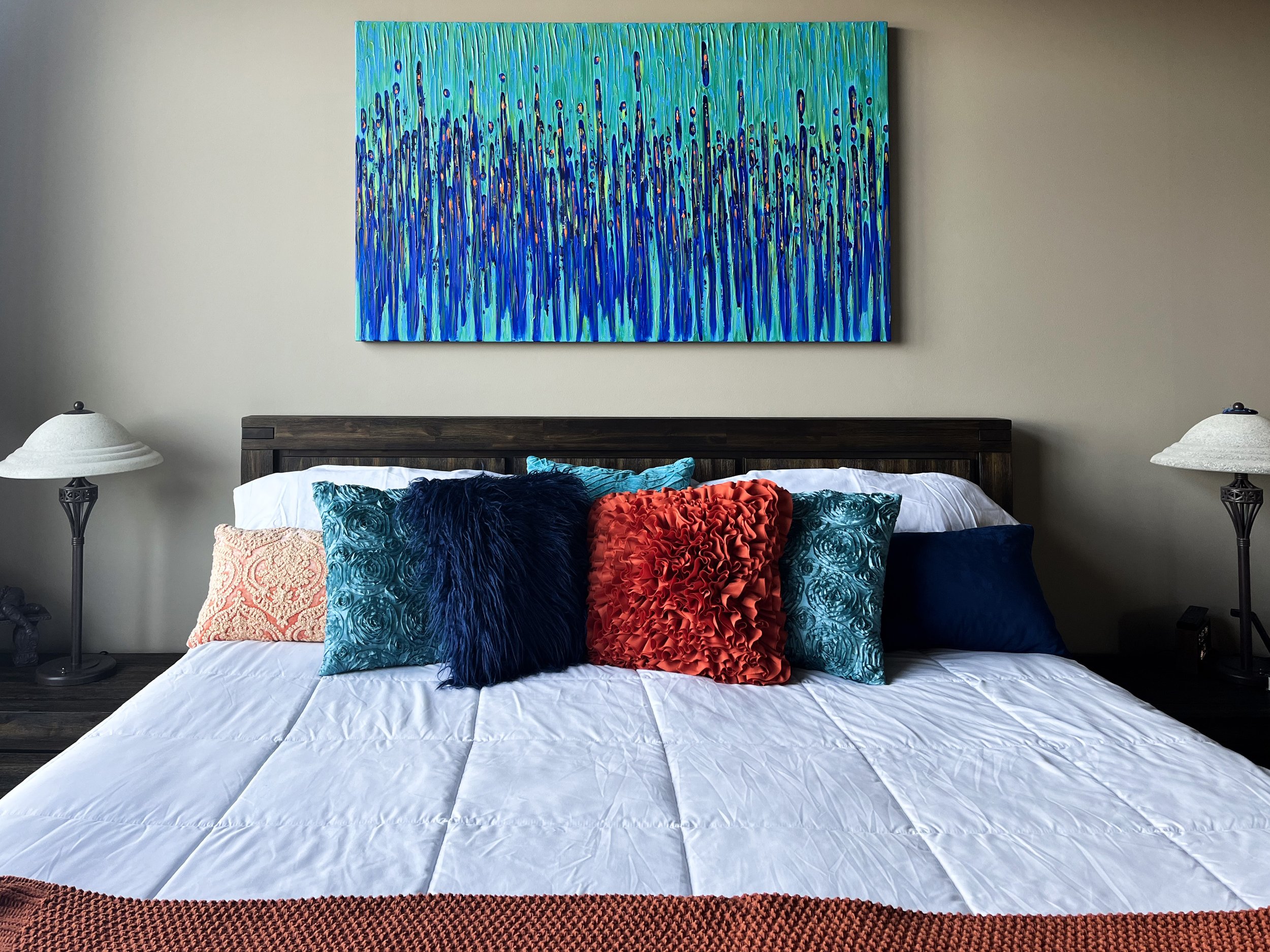A neatly made bed with a dark wooden headboard, a white comforter, and multiple decorative pillows in blue, red, and peach colors. Two matching lamps are on bedside tables on either side, and a large colorful abstract painting hangs above the bed.