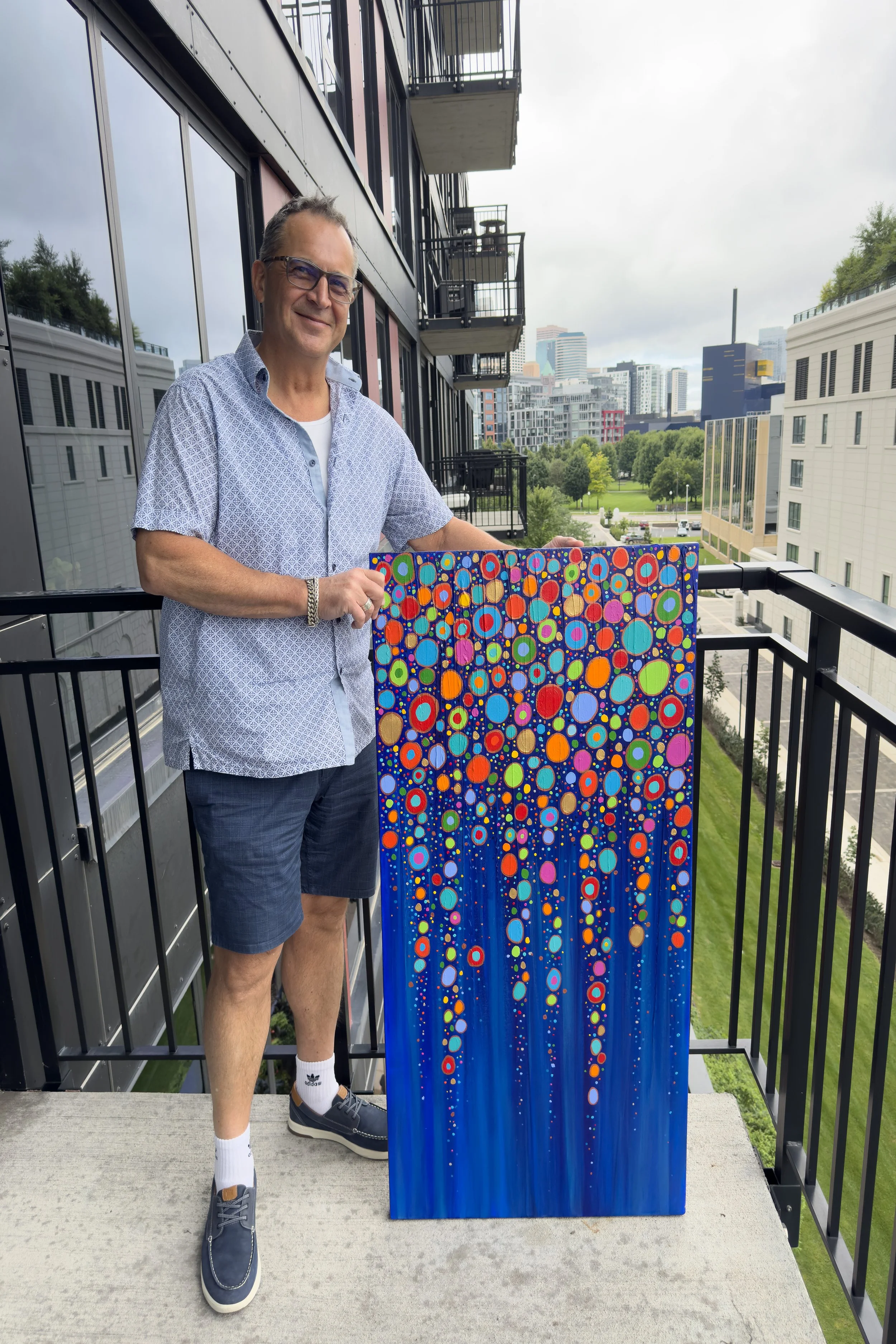 A man outdoors on a balcony holding an abstract colorful painting with circular patterns on a blue background, cityscape in the background.