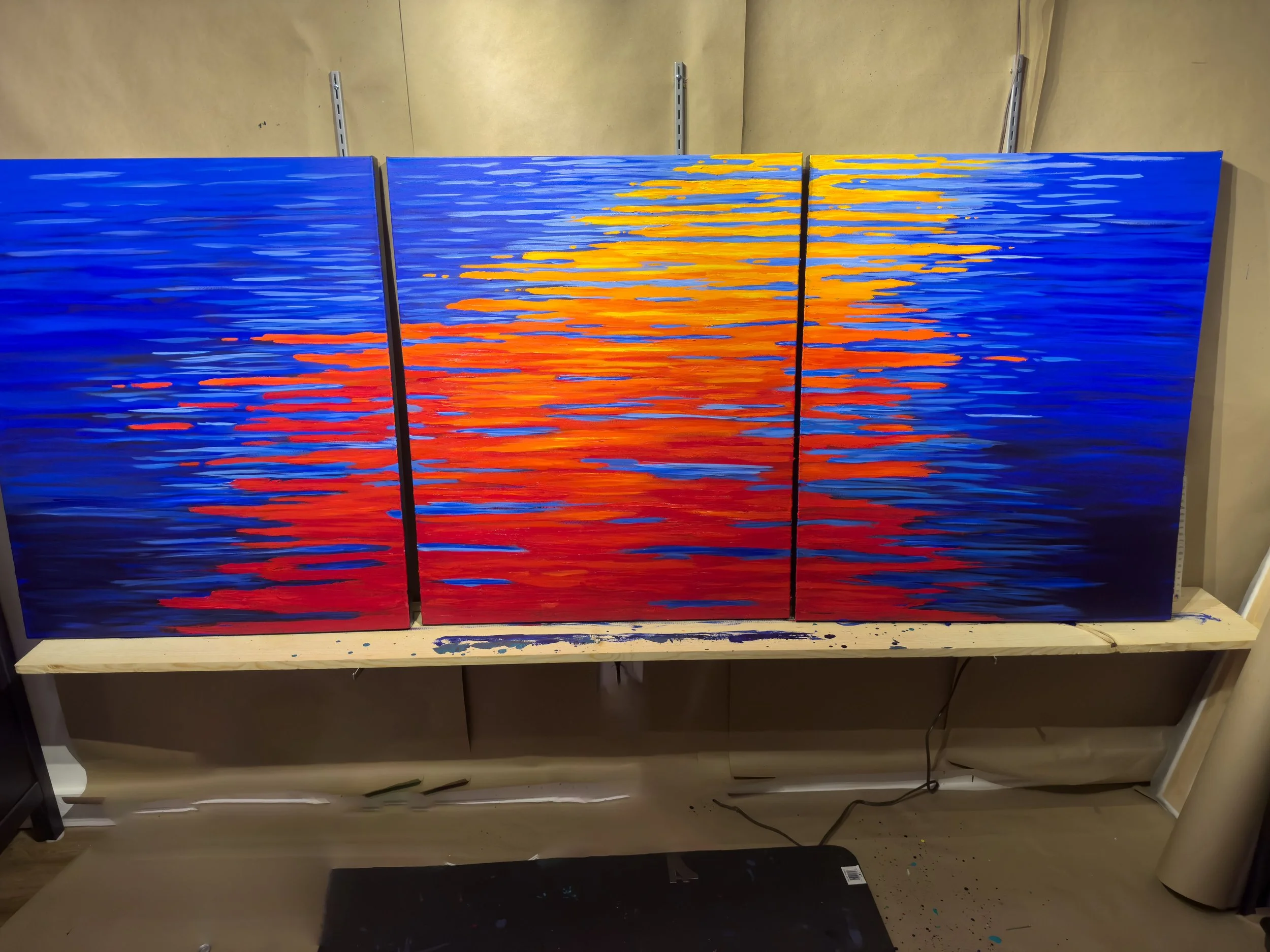 Triptych painting of a vibrant sunset over water with blue, red, orange, and yellow horizontal brushstrokes.