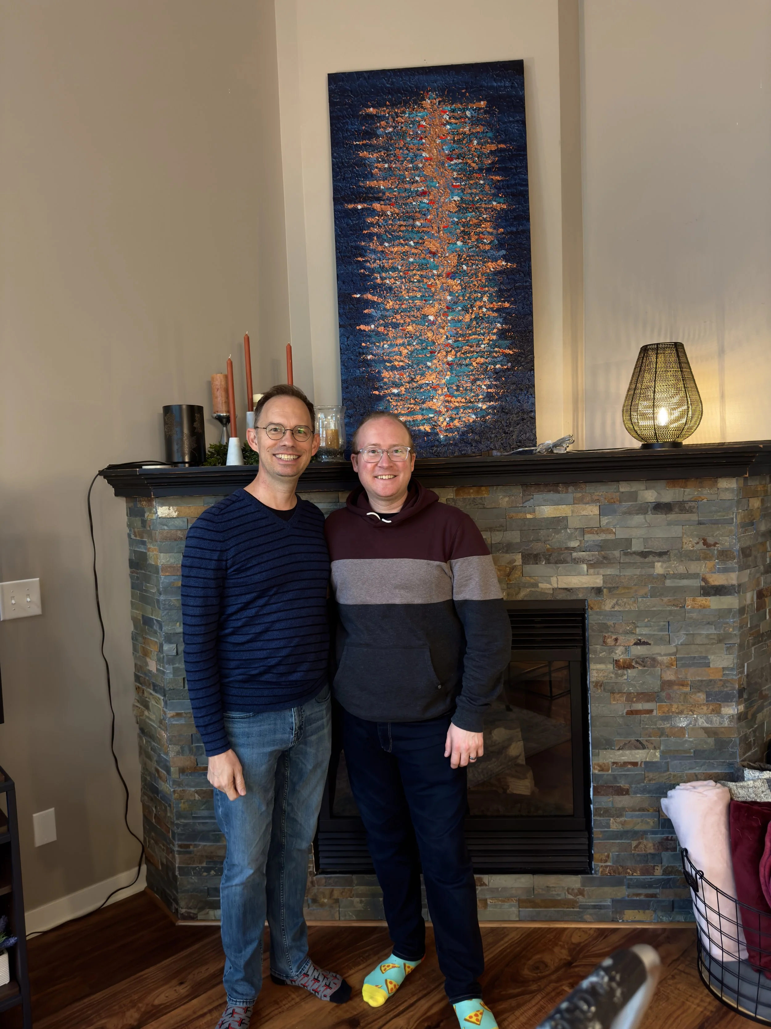 Two men smiling and standing together in front of a stone fireplace in a living room, with a large colorful abstract painting above the mantel, decorative candles, and a lamp on the mantle.