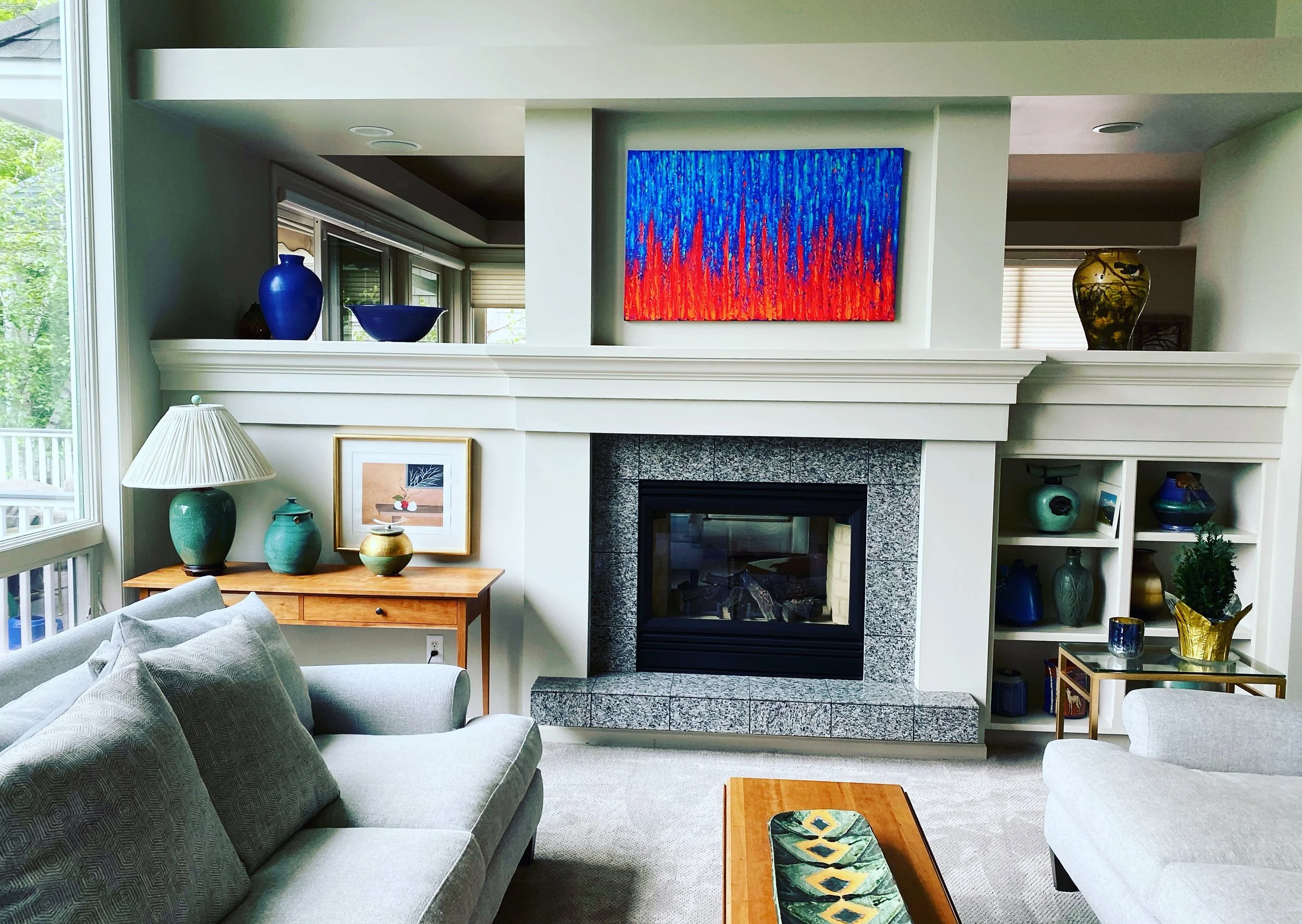 Living room with a fireplace, built-in shelves with decorative vases and objects, a framed artwork, and a colorful abstract painting above the mantel. There are two light-colored sofas, a wooden side table with a lamp and decorative vases, and a wind