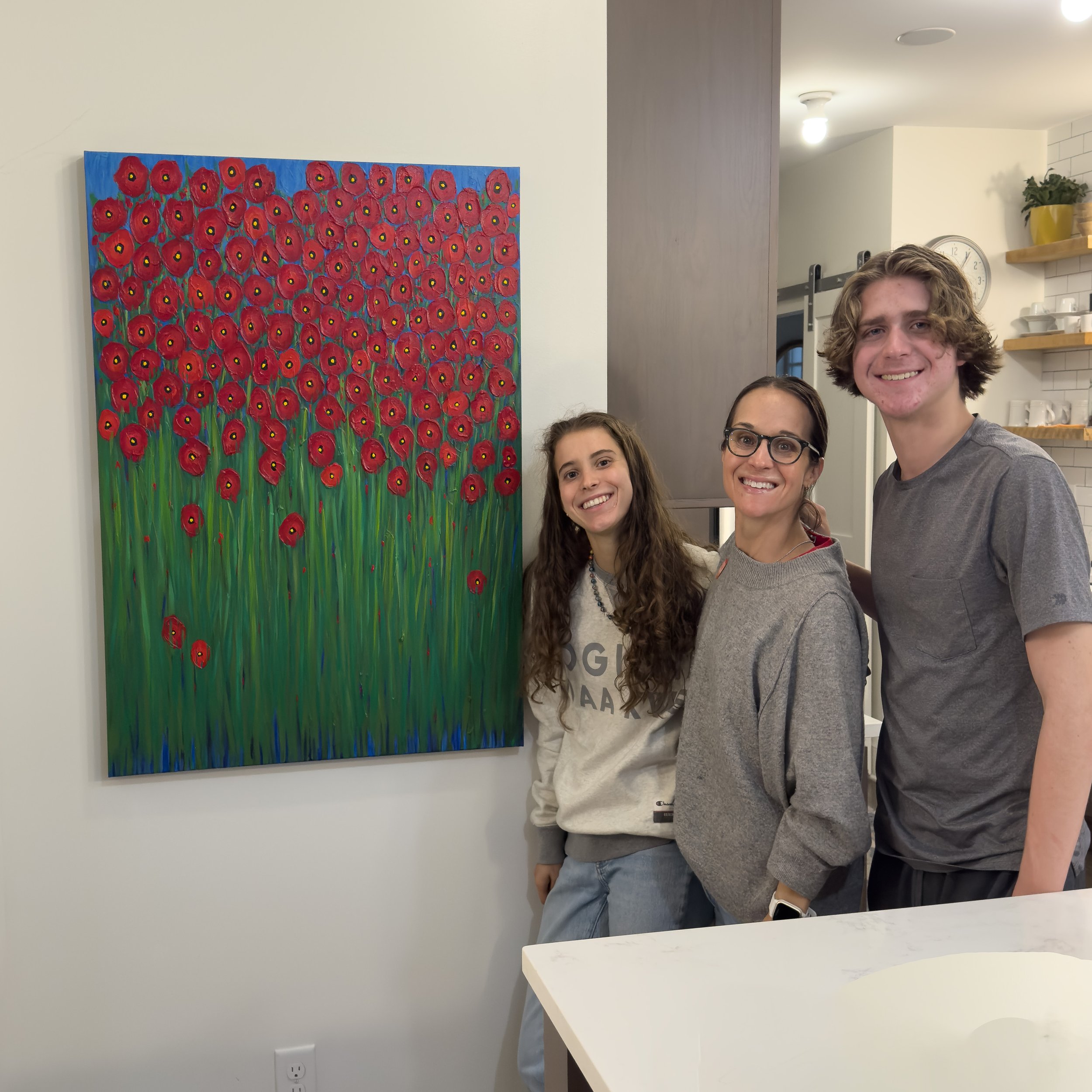 Three people smiling and standing next to a colorful painting of numerous red poppies on a green background inside a room.