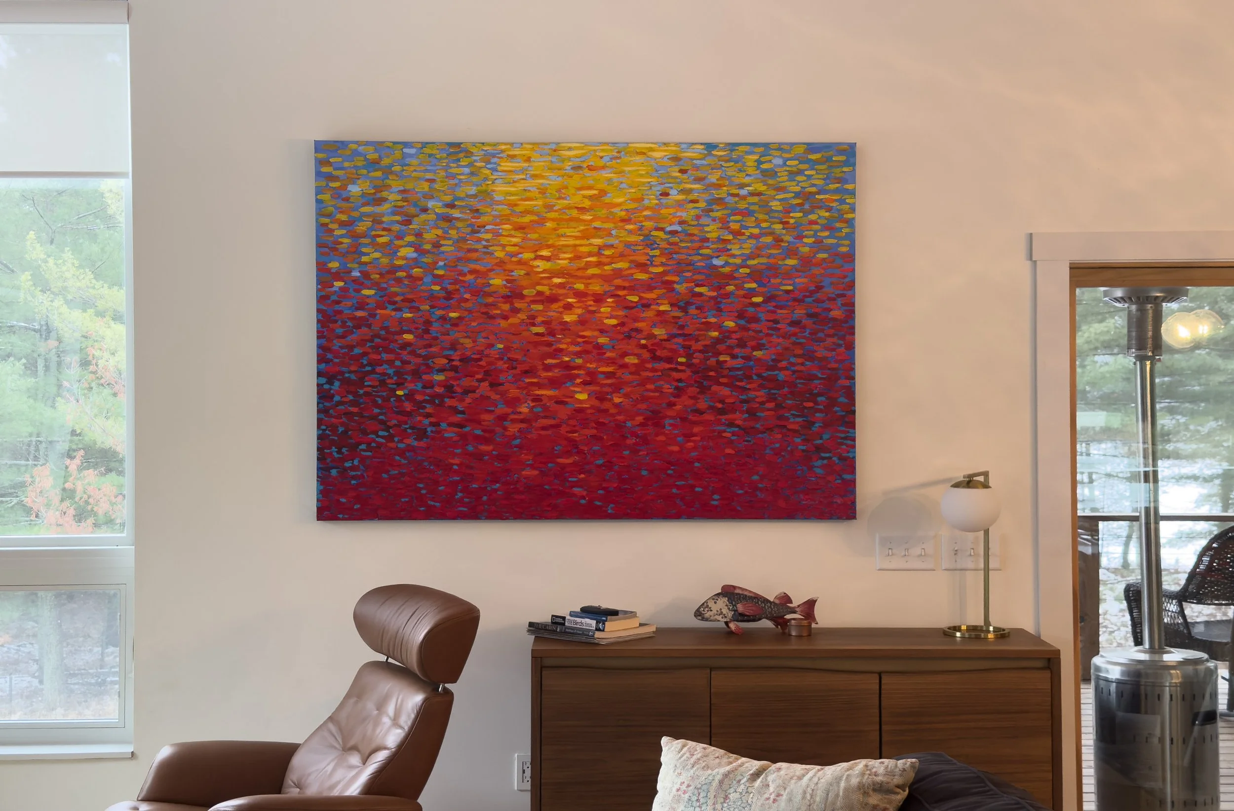 A colorful abstract painting mounted on a white wall, with a mix of red, yellow, blue, and orange dot patterns resembling a sunset or sunrise reflection.