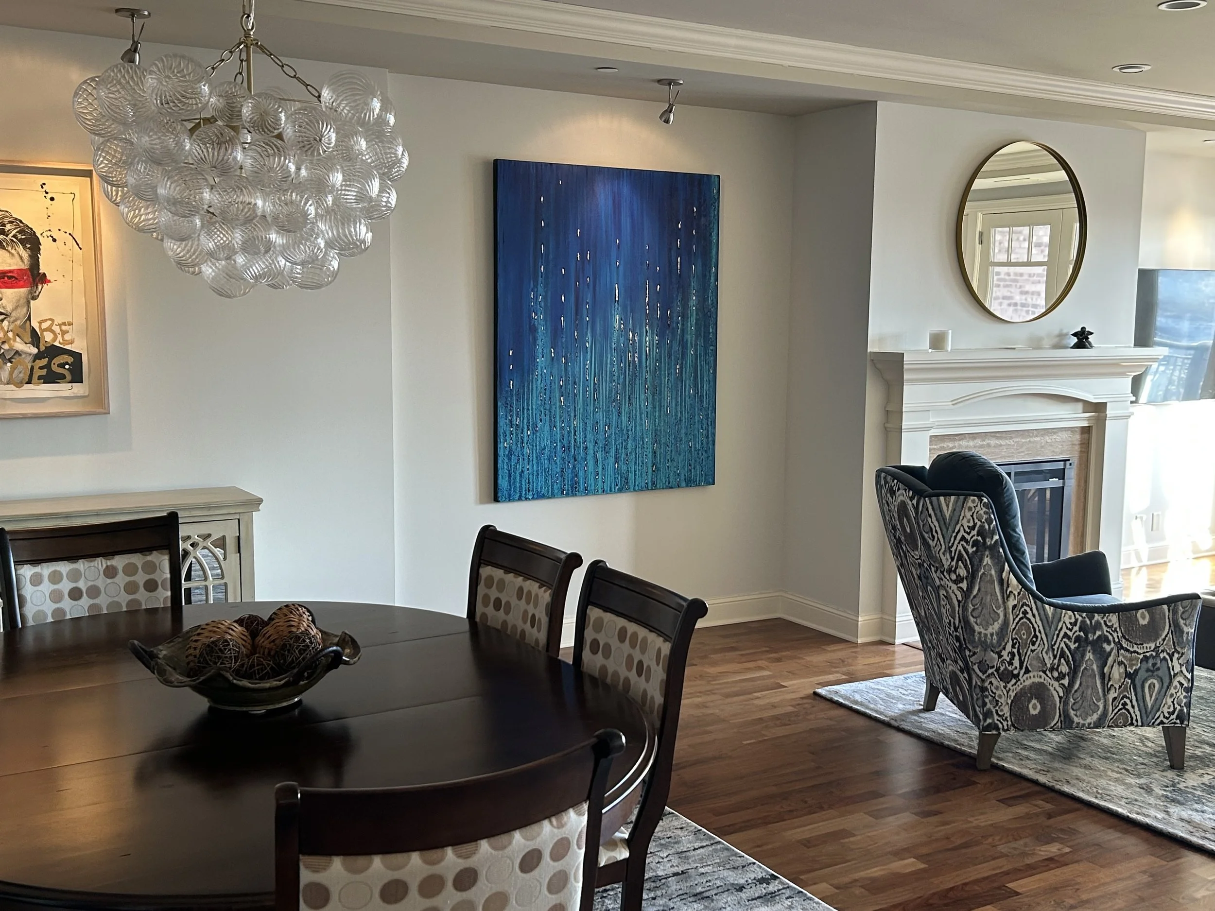 A living room with a white fireplace, a patterned armchair, a blue abstract painting, a mirror above the fireplace, a round dining table with chairs, a decorative bowl with balls on the table, and a white chandelier hanging from the ceiling.