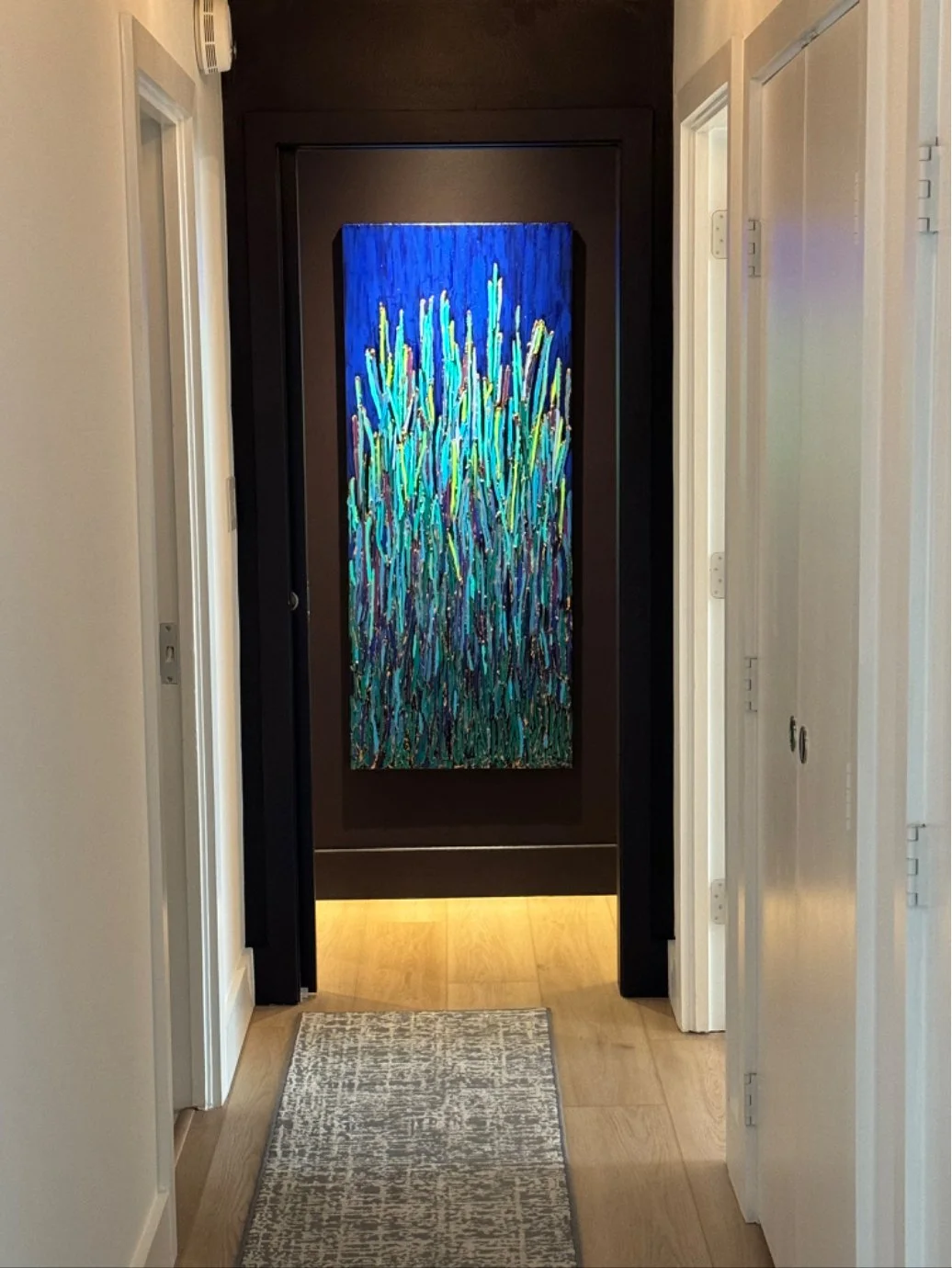 A hallway with a colorful abstract painting of tall grass behind a black frame on the black wall at the end of the hall. The hall has white walls and doors, a light-colored wooden floor, and a small patterned rug.