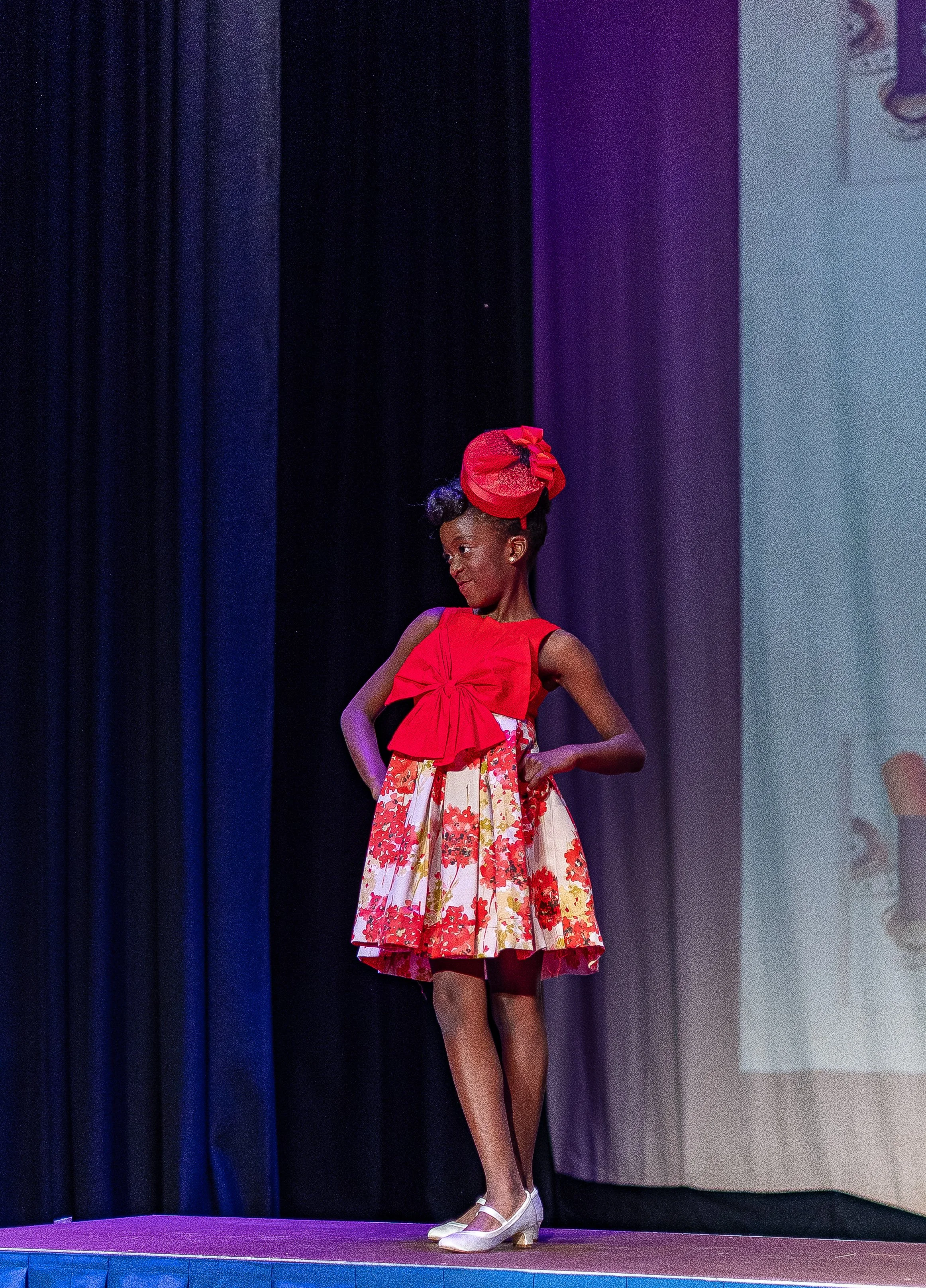 Young girl on stage wearing a red dress with a floral skirt, red bow on her head, and white shoes, posing confidently during a performance.