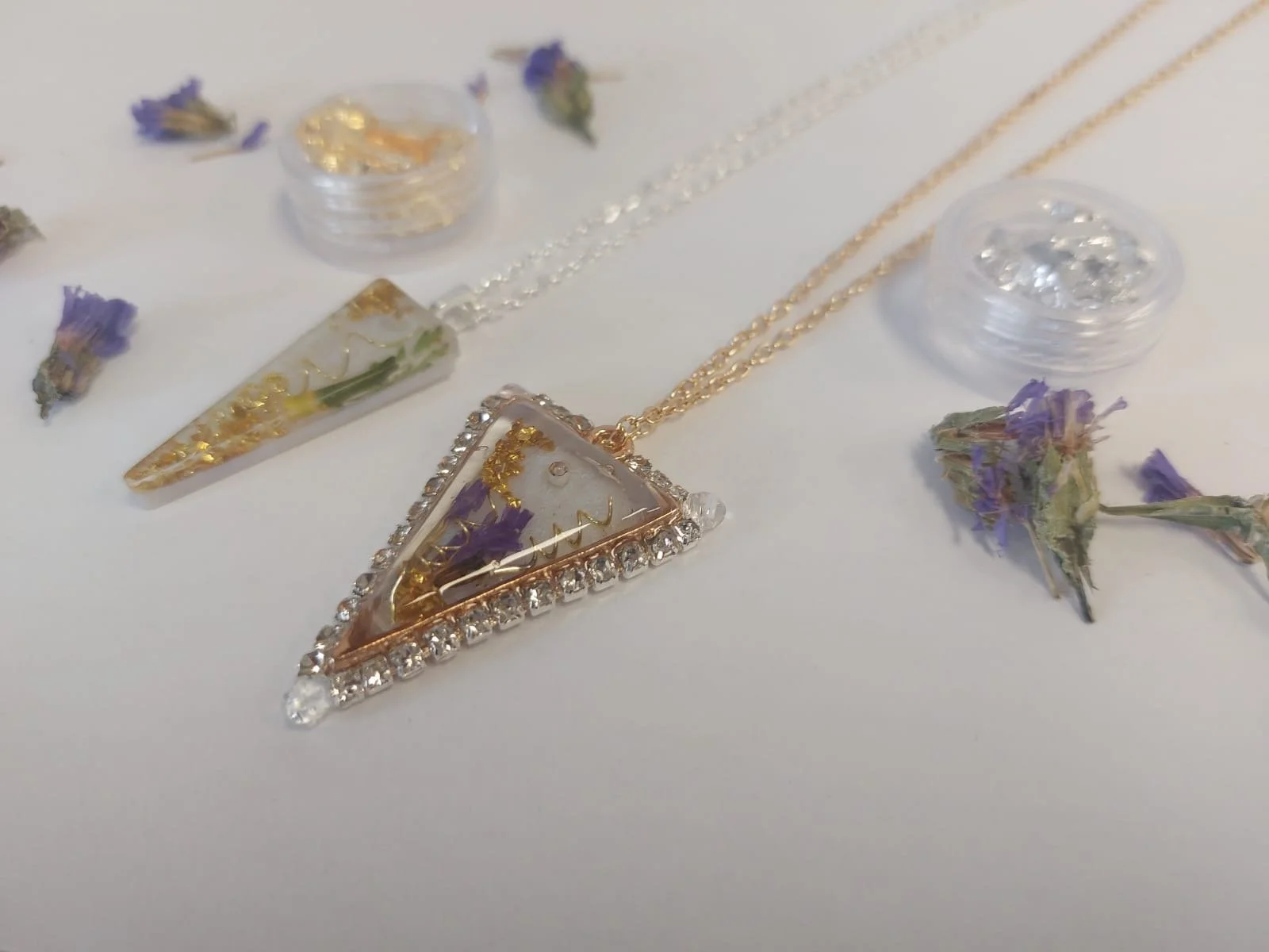 Close-up of jewelry including a triangular pendant with embedded flowers and rhinestones, a triangular-shaped earring with jewels, and gold chains, surrounded by dried purple flowers and small plastic containers of rhinestones on a white surface.