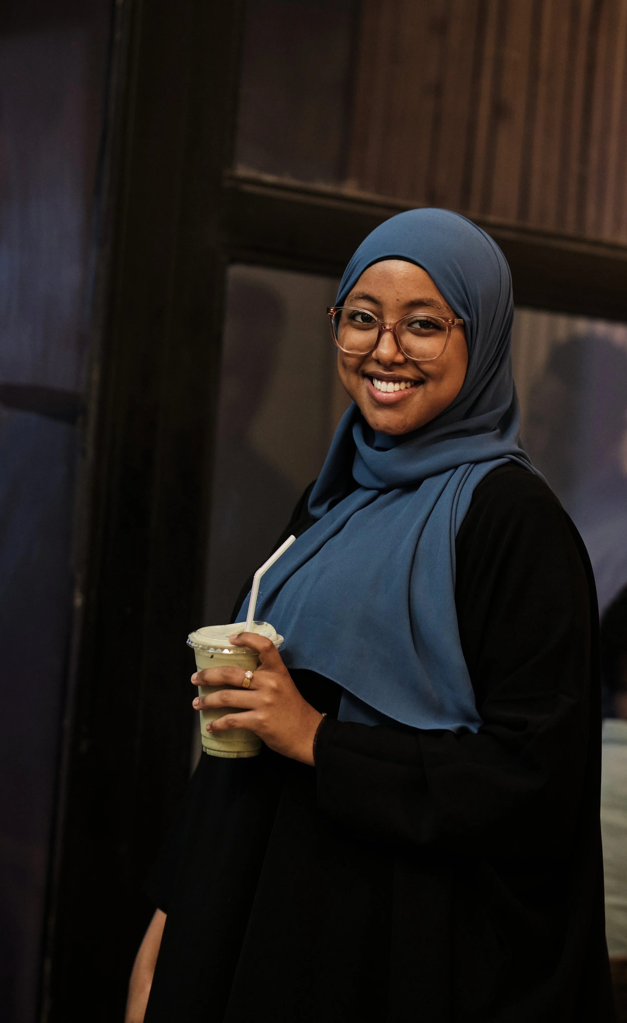 A smiling woman wearing a blue headscarf and glasses holding a drink with a straw in a dark indoor setting.