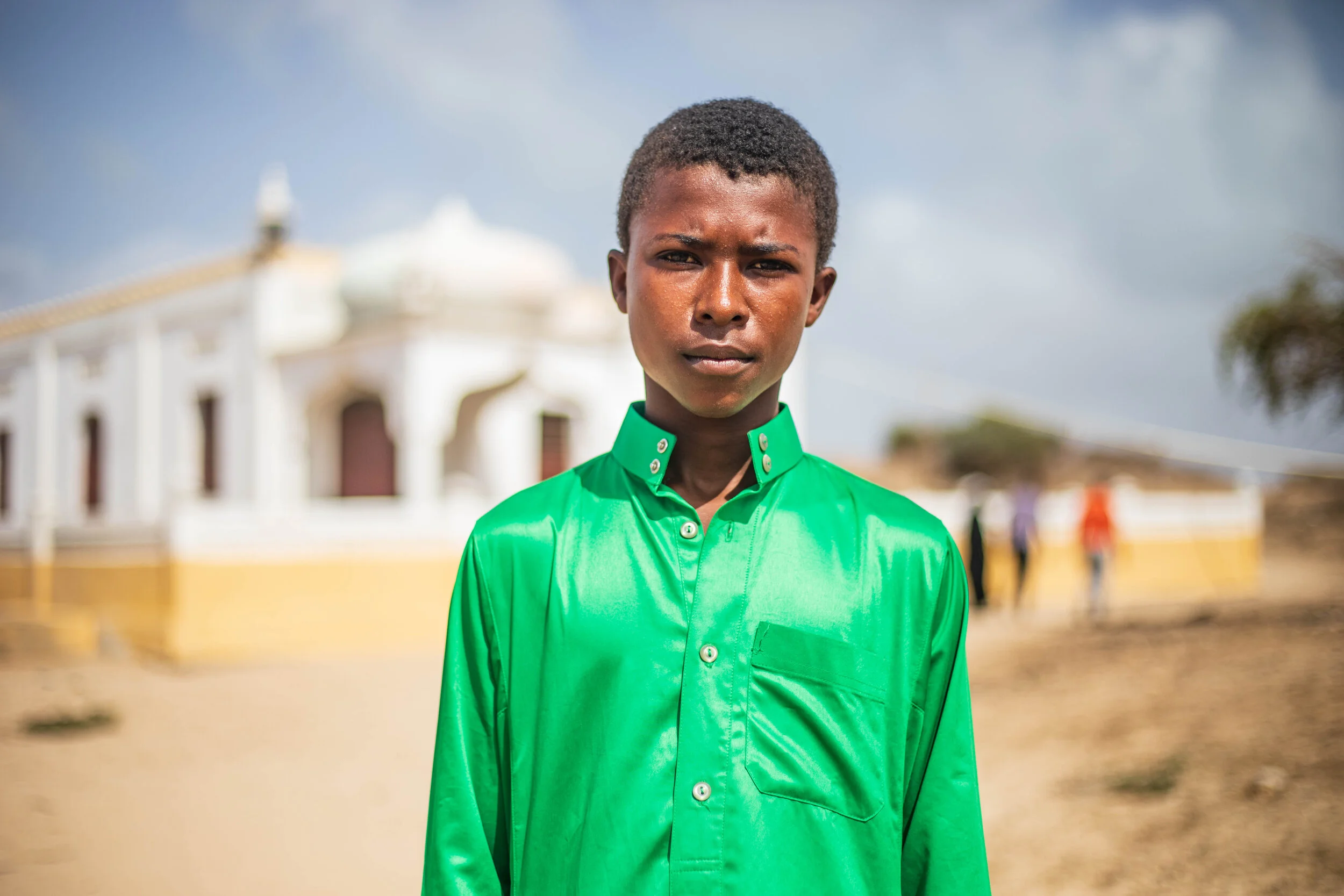 A young man with short, curly black hair and dark skin wearing a shiny green shirt standing outdoors on a sunny day.