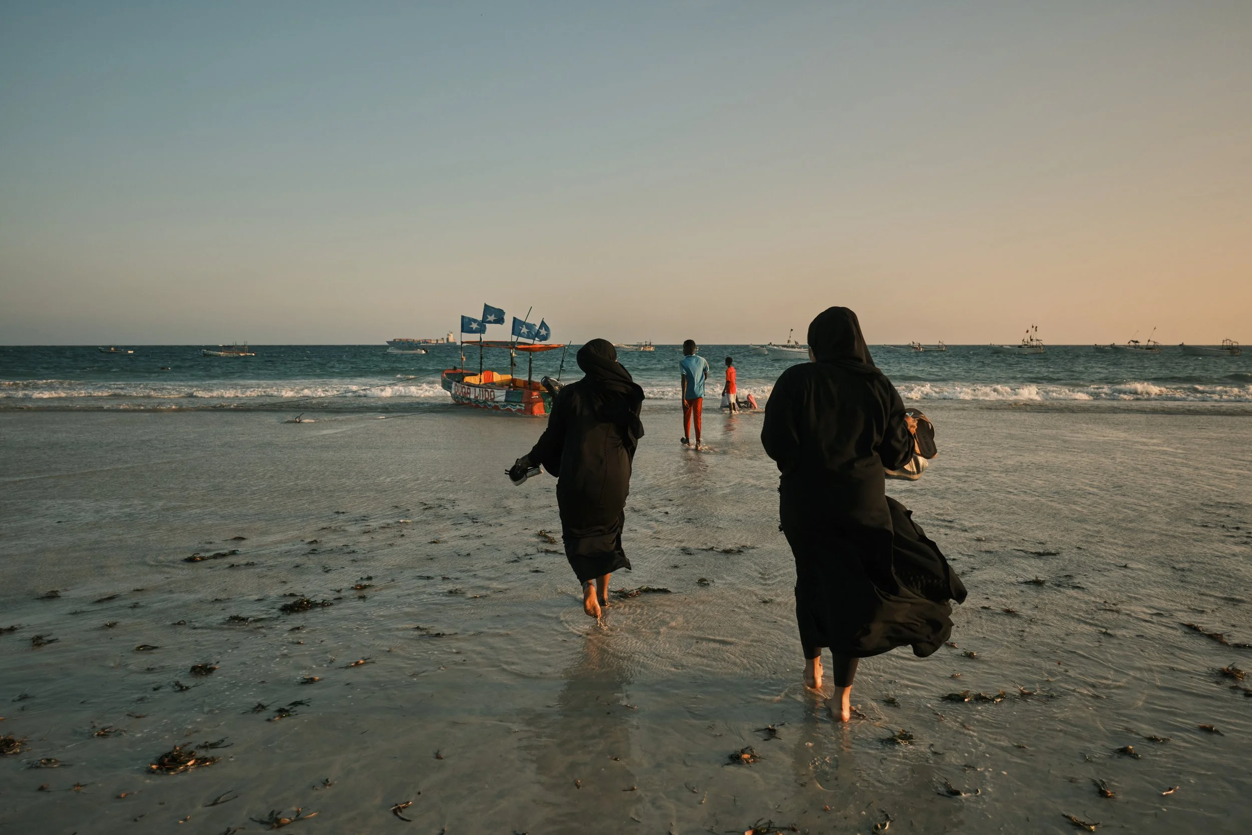 People walking on a beach with waves, boats in the water, and a boat with French flags in the background.