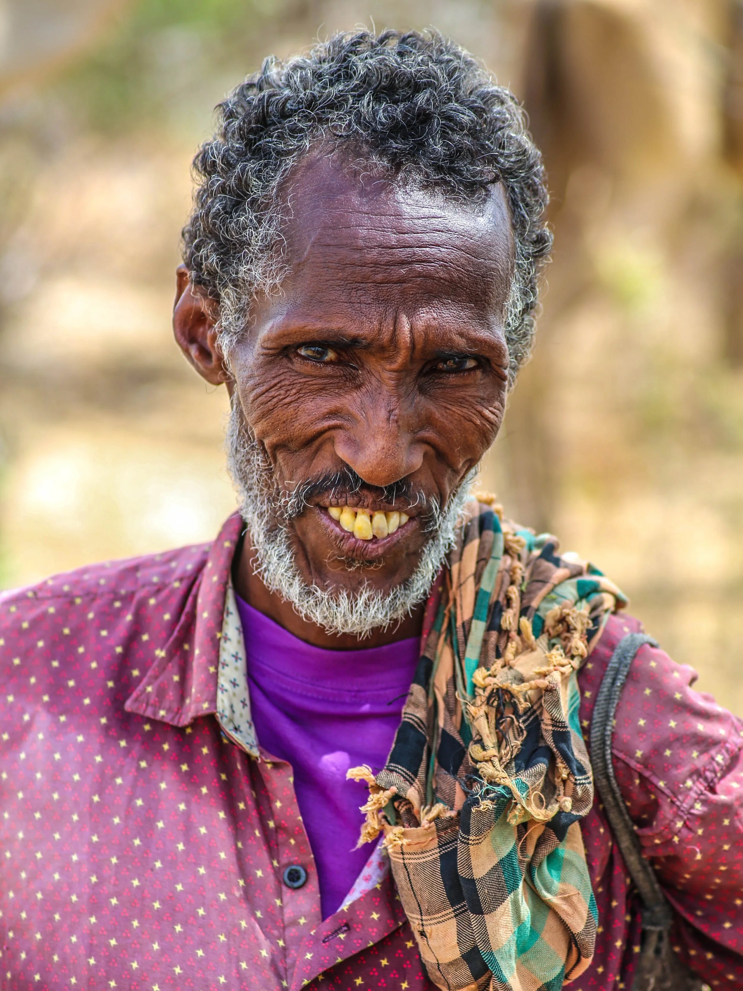 Close-up of an elderly man with gray curly hair, showing a big smile with yellowed teeth, wearing a purple shirt and a red jacket with a star pattern, outdoors with a blurred natural background.
