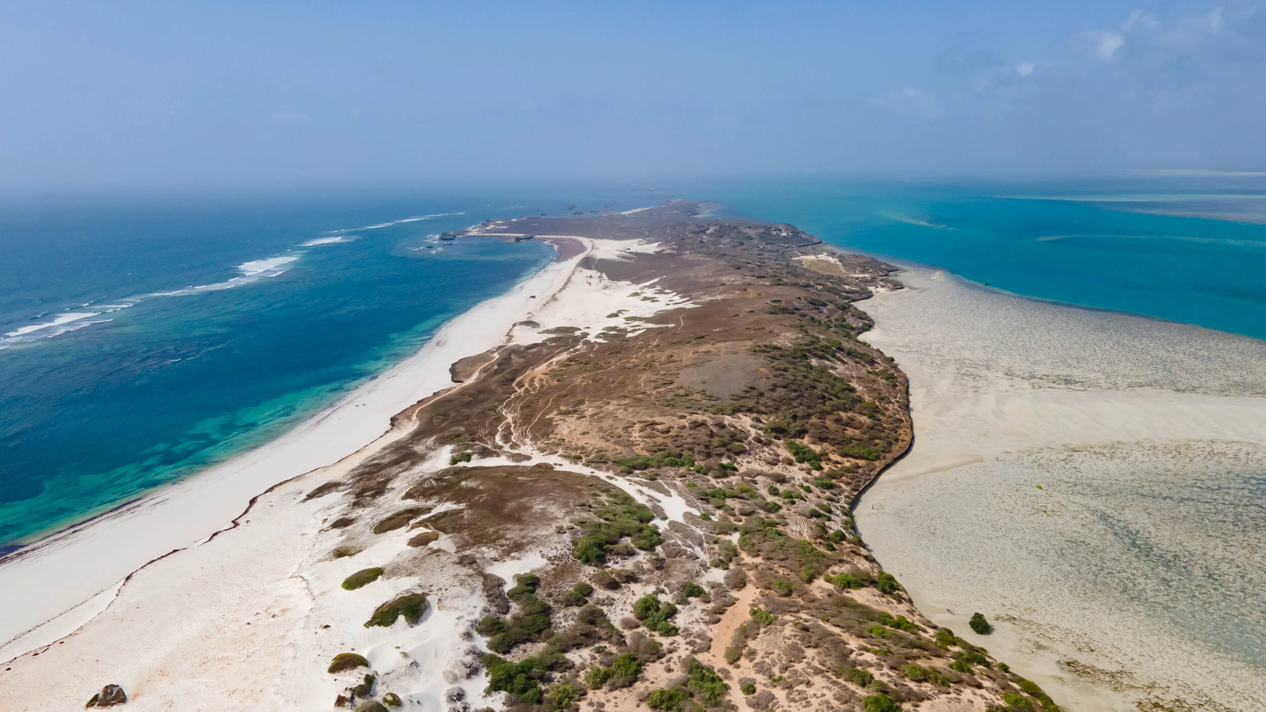 Aerial view of a sandy peninsula with patches of vegetation, surrounded by turquoise ocean water and white beaches.
