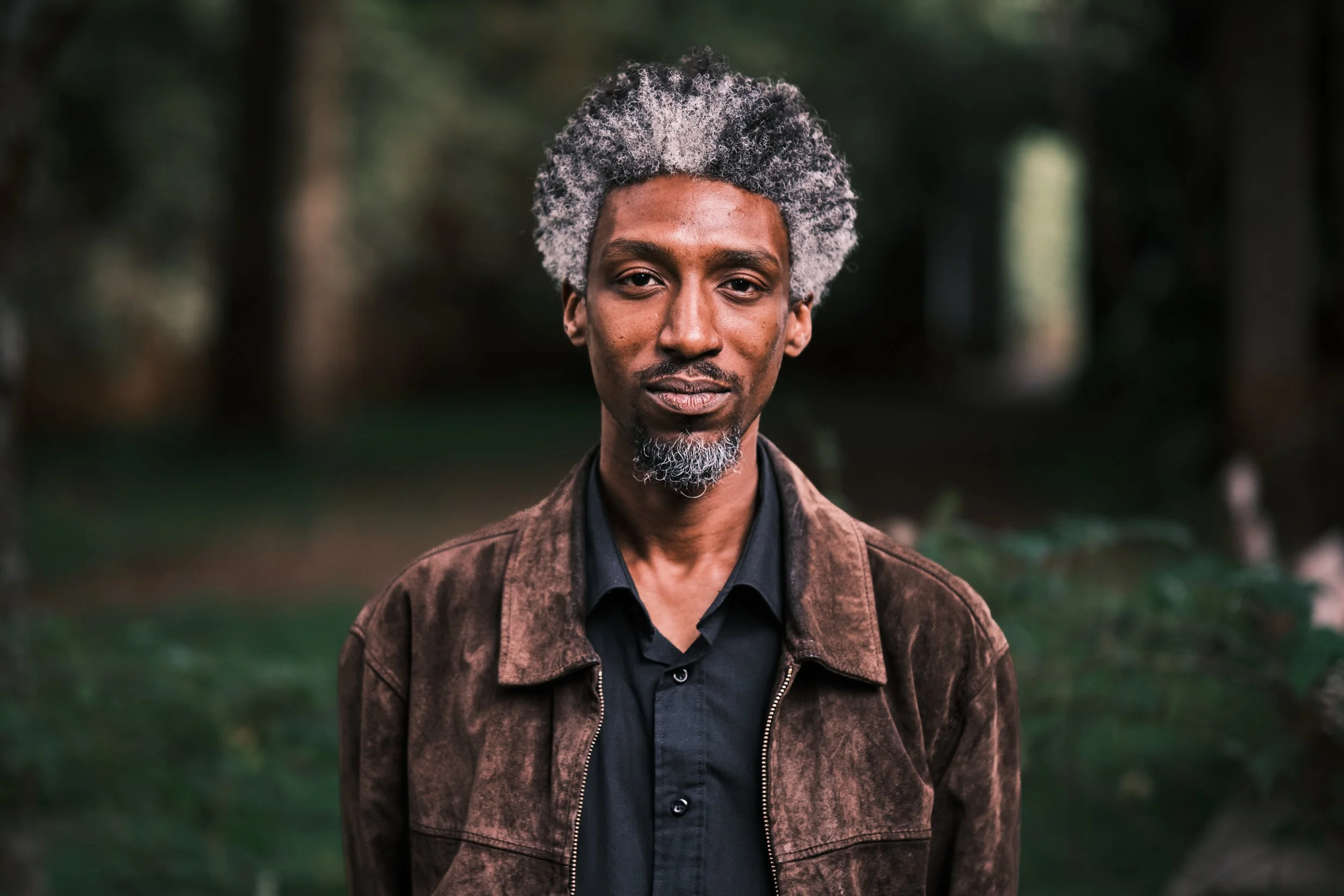 Portrait of a man with gray and black hair, wearing a black shirt and a brown jacket, standing outdoors with blurred trees in the background.