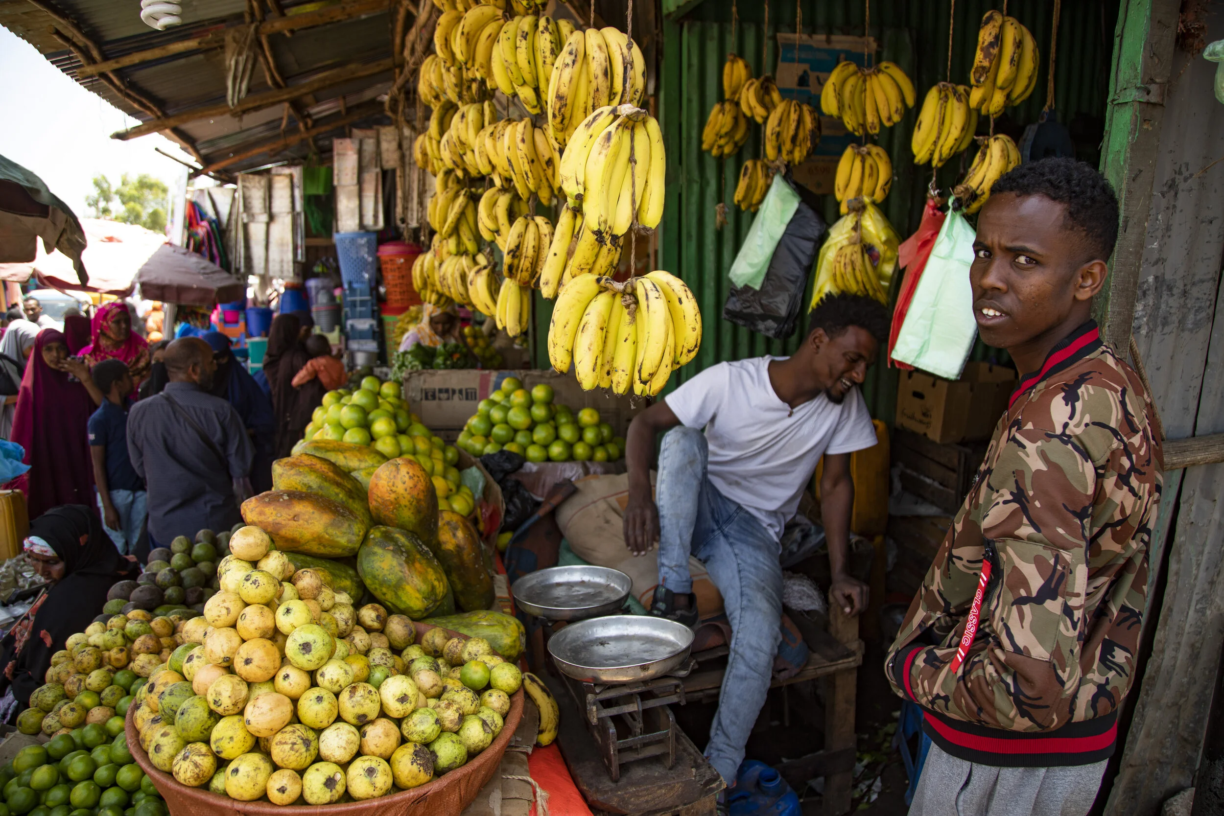A bustling outdoor market with bananas hanging from above, and various fruits like tomatoes and papayas displayed on stalls. Several vendors and shoppers are engaged in discussions amid colorful umbrellas and a lively crowd.