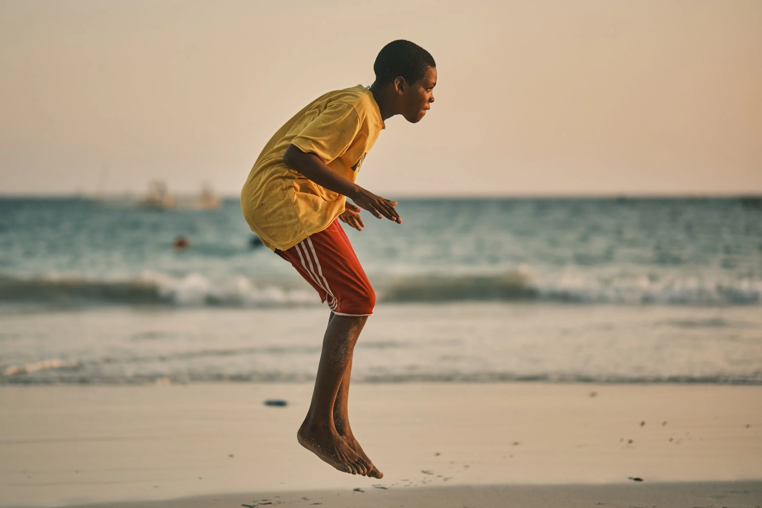 A young person in a yellow shirt and red shorts is jumping on the beach near the ocean, with their feet low to the water and their body bent forward, appearing to be playing or jumping.