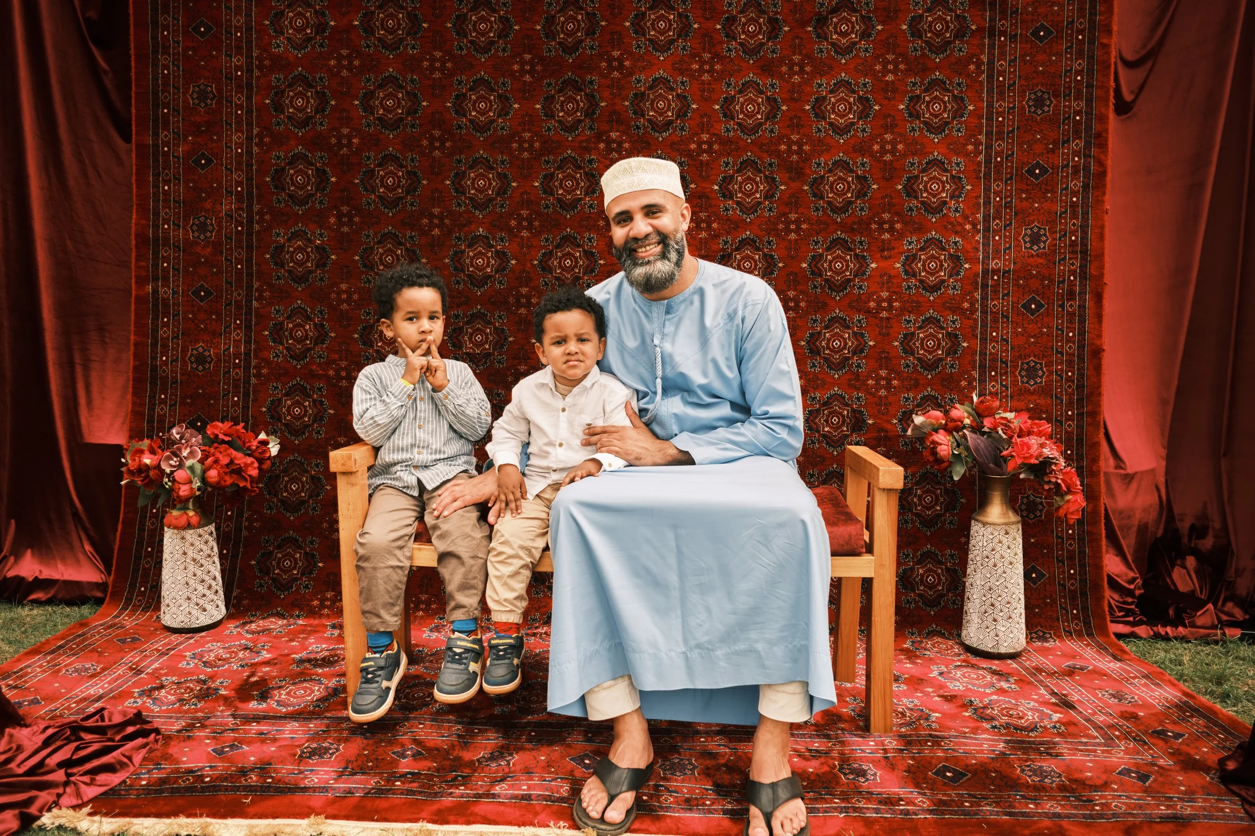 A man with a beard and white prayer cap sitting on a wooden throne, flanked by two young boys, under a red patterned rug backdrop, with floral arrangements on either side.