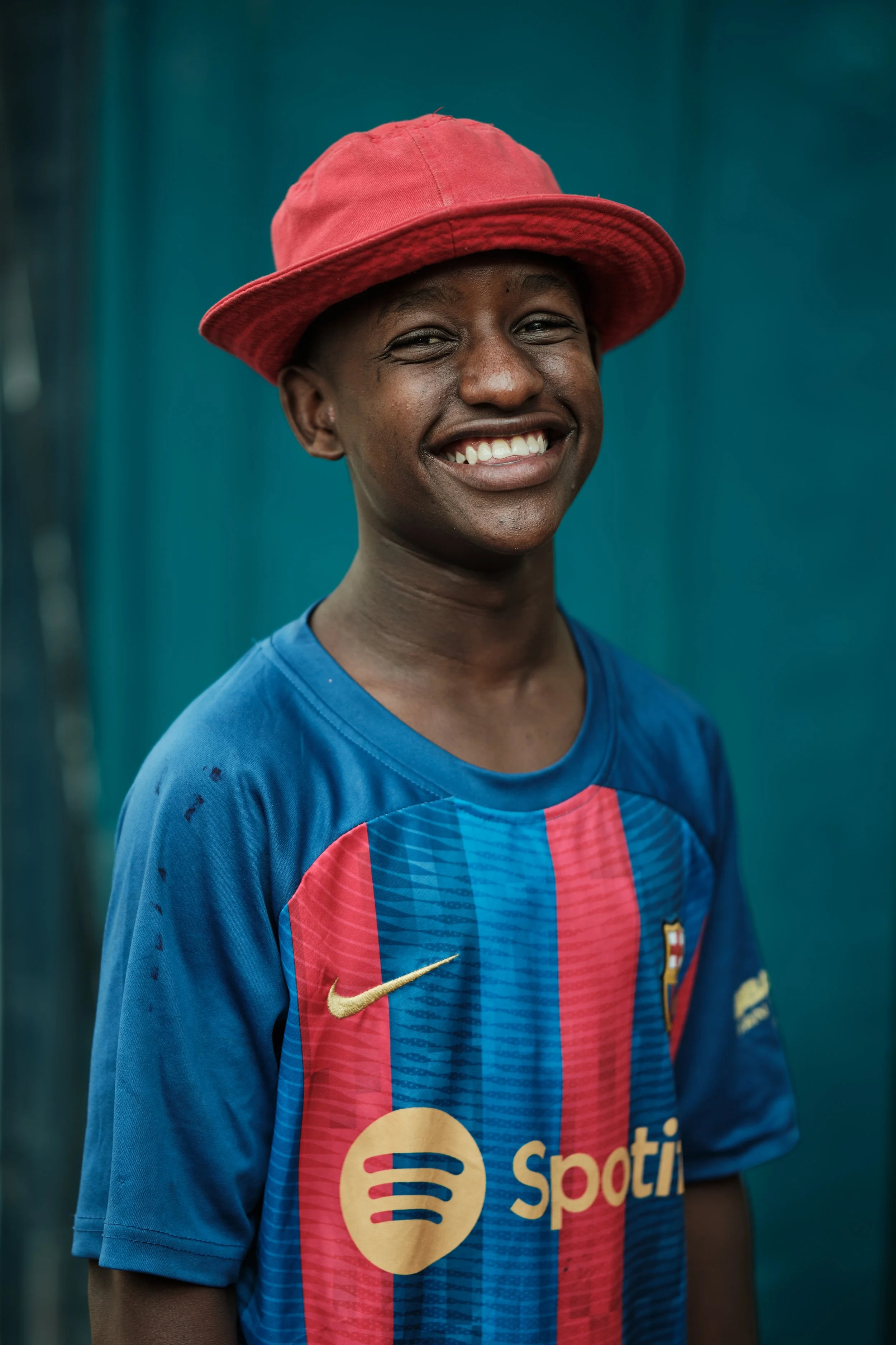 A young boy with dark skin and a broad smile, wearing a red hat and a blue and red sports jersey with the Spotify logo.