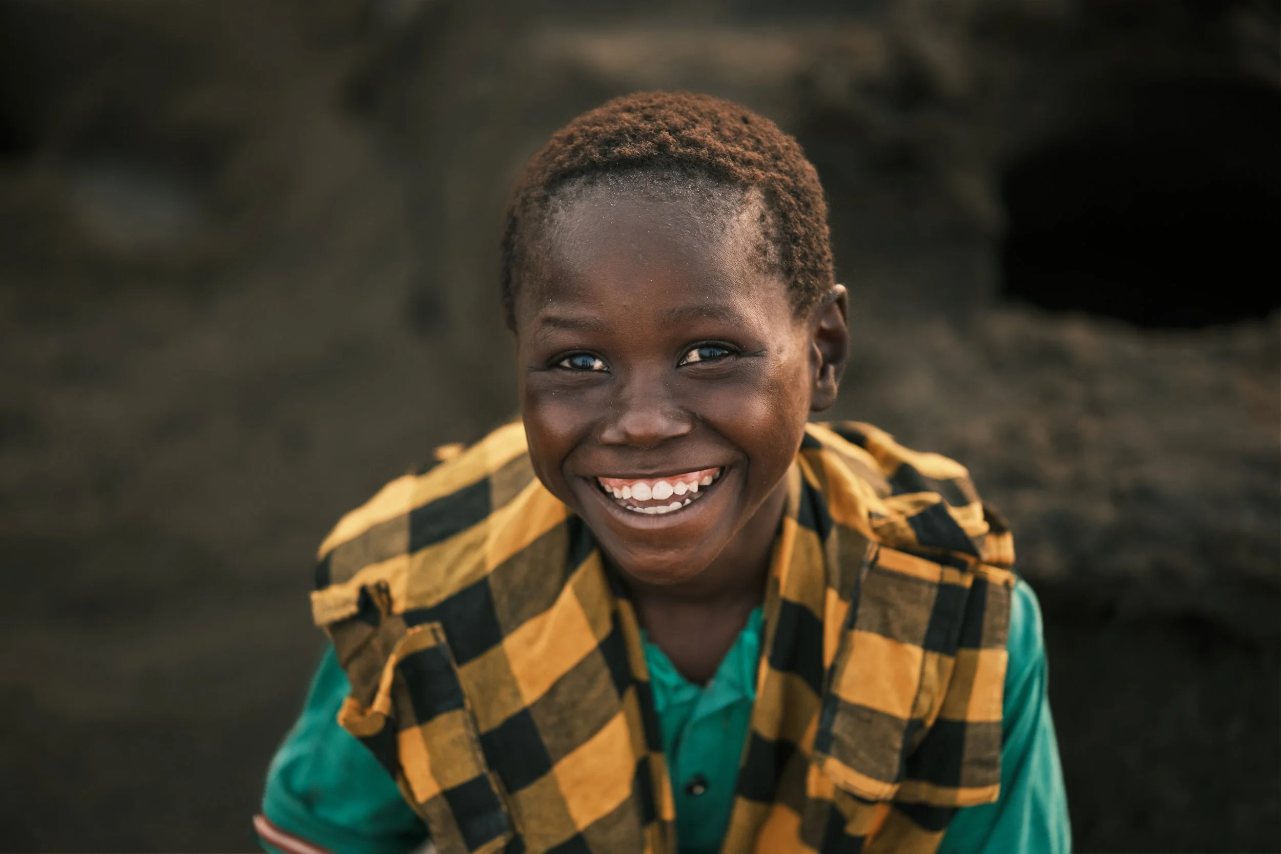 A close-up of a smiling young boy with short, textured hair, wearing a yellow and black checkered jacket over a teal shirt, outdoors with a blurry dark background.