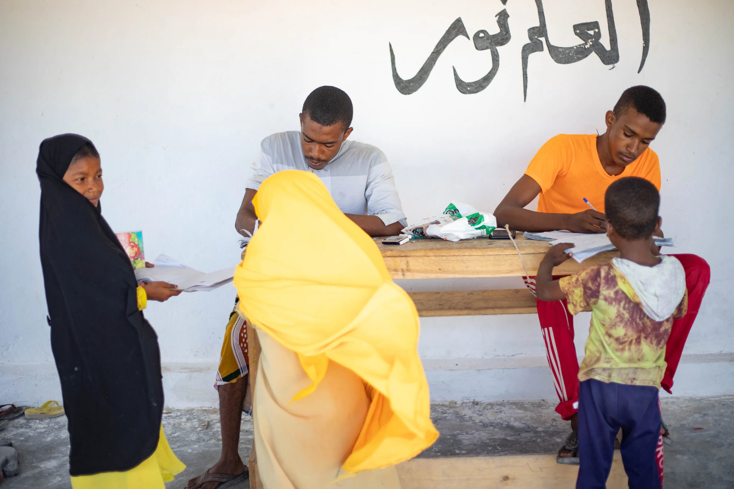 Teachers check students assignments in Mudawa Island School. 

This school is the only one on the island. There is an Arabic phrase written on the wall behind them which reads “Knowledge is light”. 