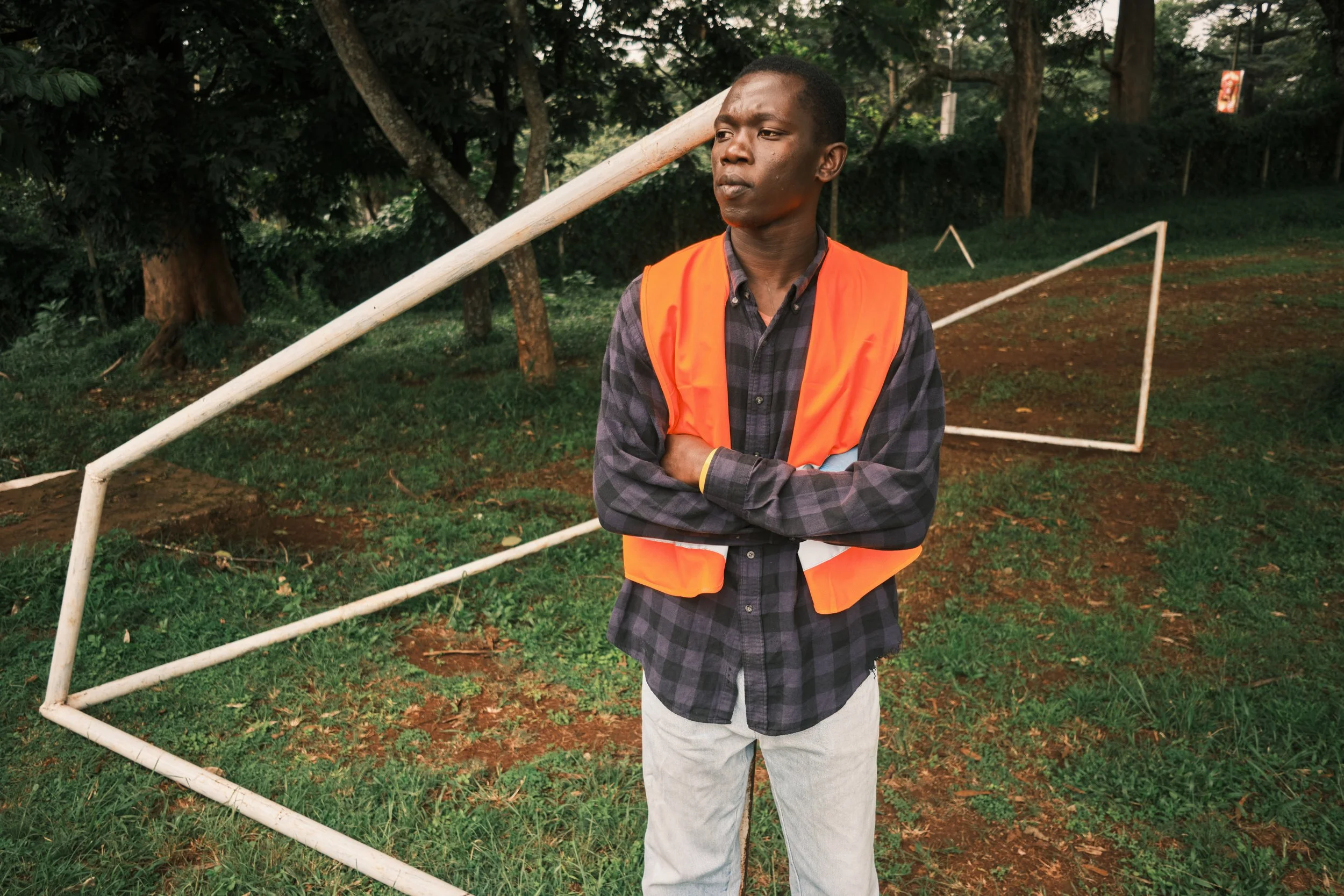 A young man wearing an orange safety vest and a plaid shirt stands outdoors with crossed arms, in front of a damaged white metal fence, surrounded by trees.