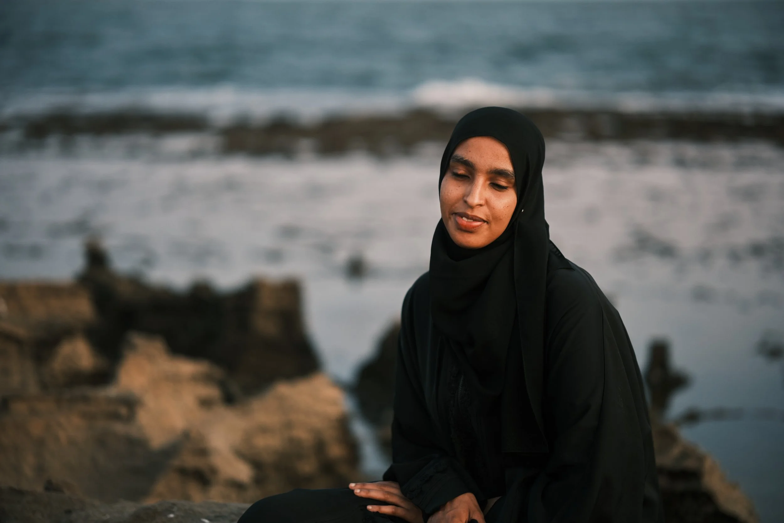 A woman wearing a black hijab sitting outdoors near the water with waves in the background.