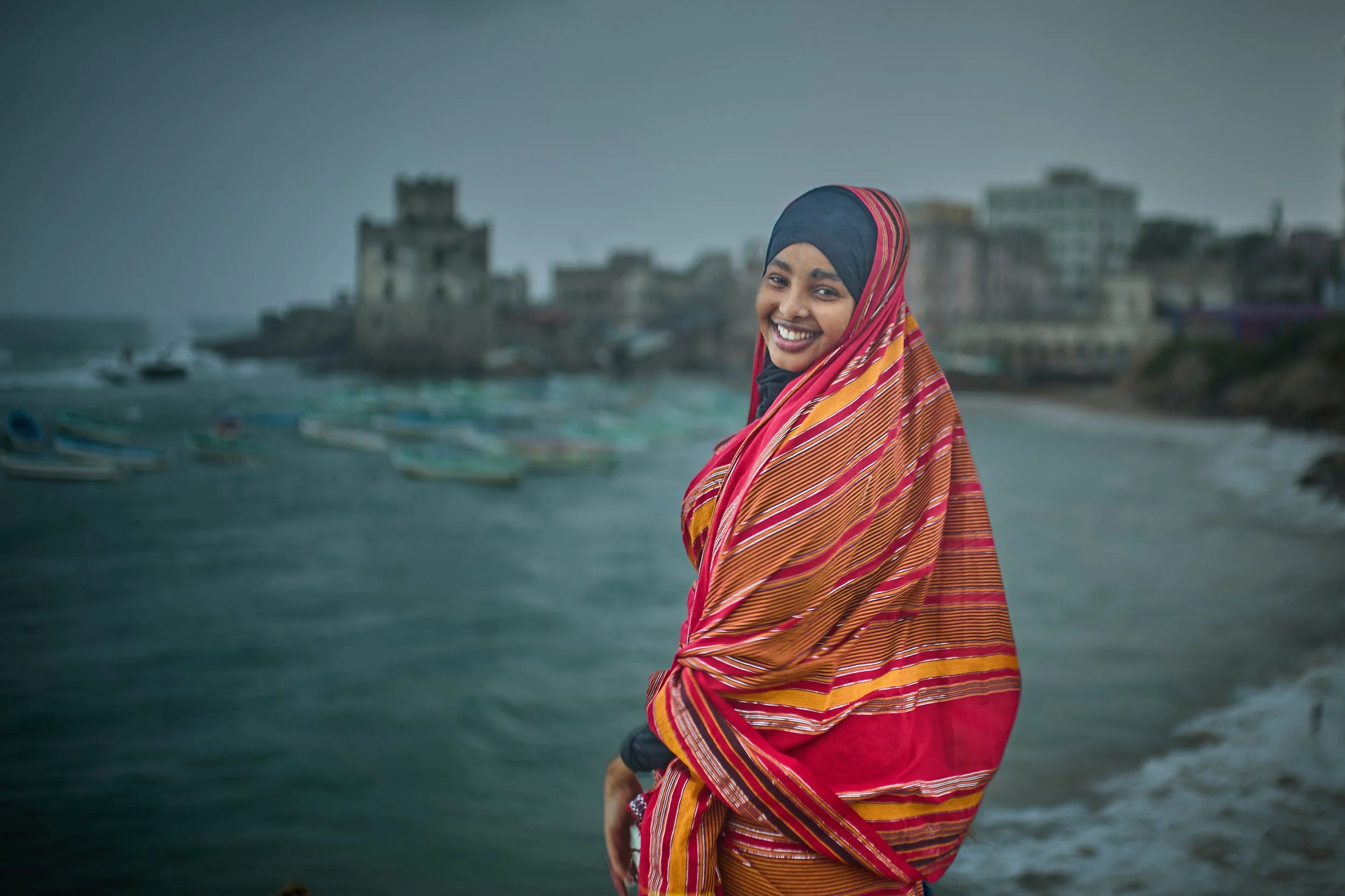 A woman wearing a colorful striped shawl and headscarf smiling at the camera with a coastal cityscape and boats in the background.