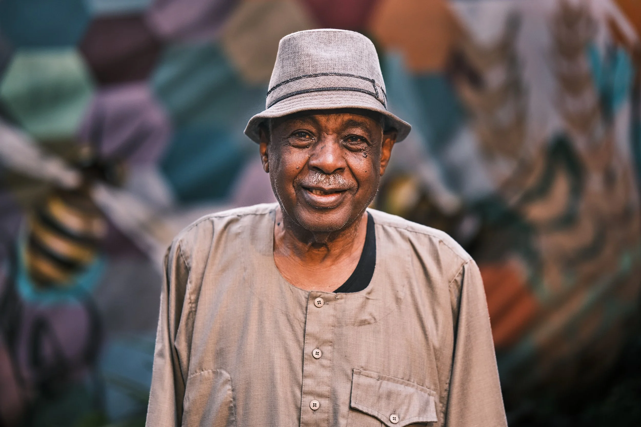 An elderly man with dark skin, wearing a gray hat with black bands and a beige shirt, smiling and standing outdoors with blurred colorful objects in the background.