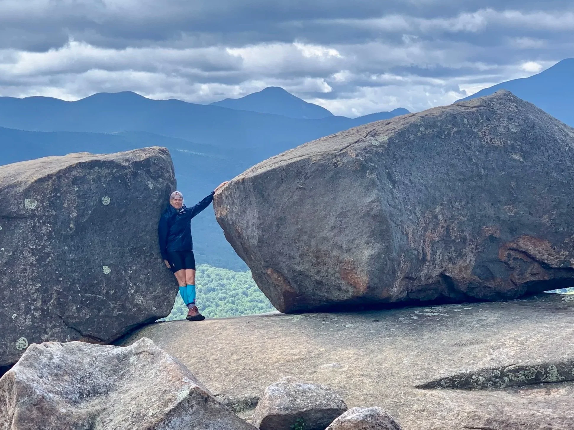 A artist hiking on a mountain with a mountain range and cloudy sky in the background.