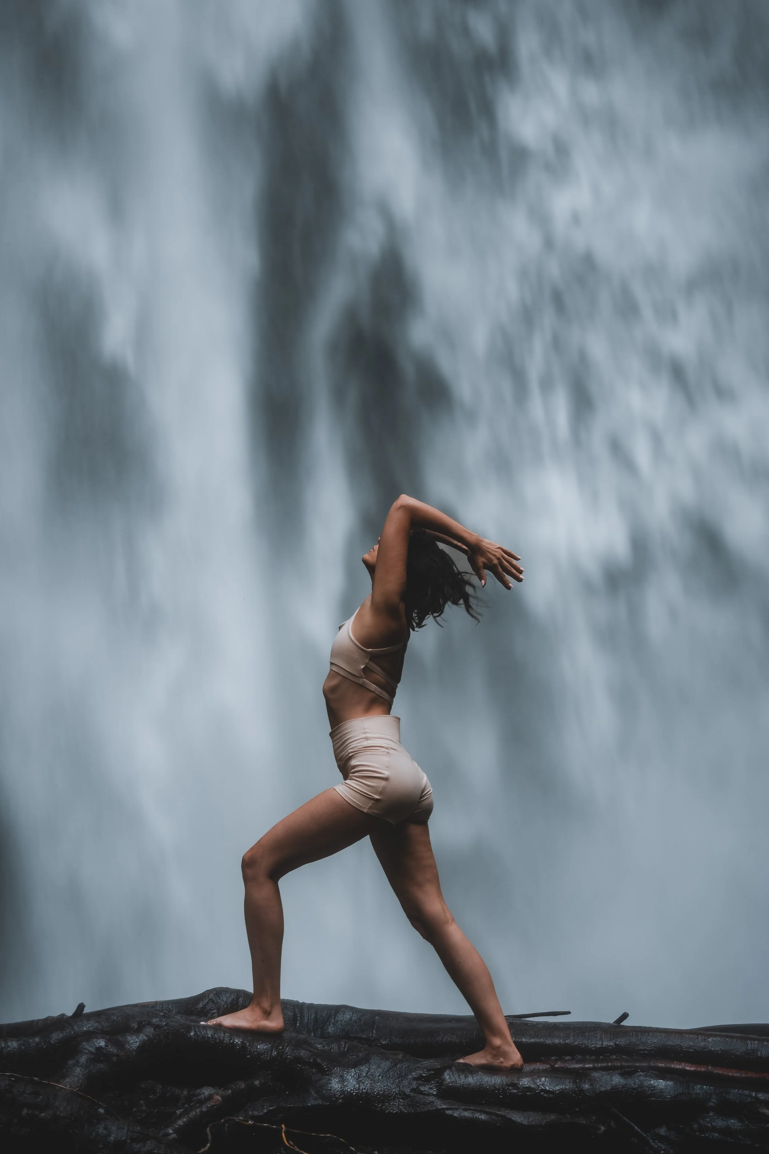 A woman in minimal clothing is balancing on a large rock in front of a waterfall, with her hands raised above her head.