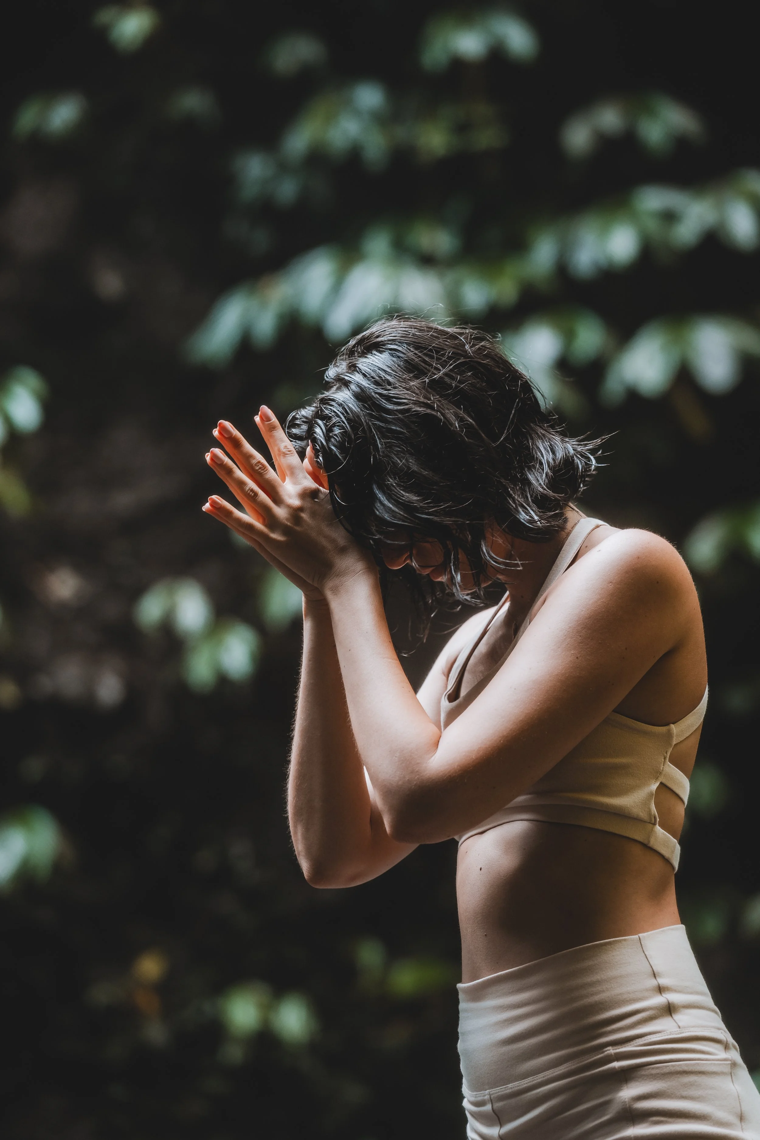 A woman with dark, wavy hair and light skin, wearing a beige sports bra and matching pants, standing outdoors with her hands covering her face, in a contemplative pose against a blurred green background.