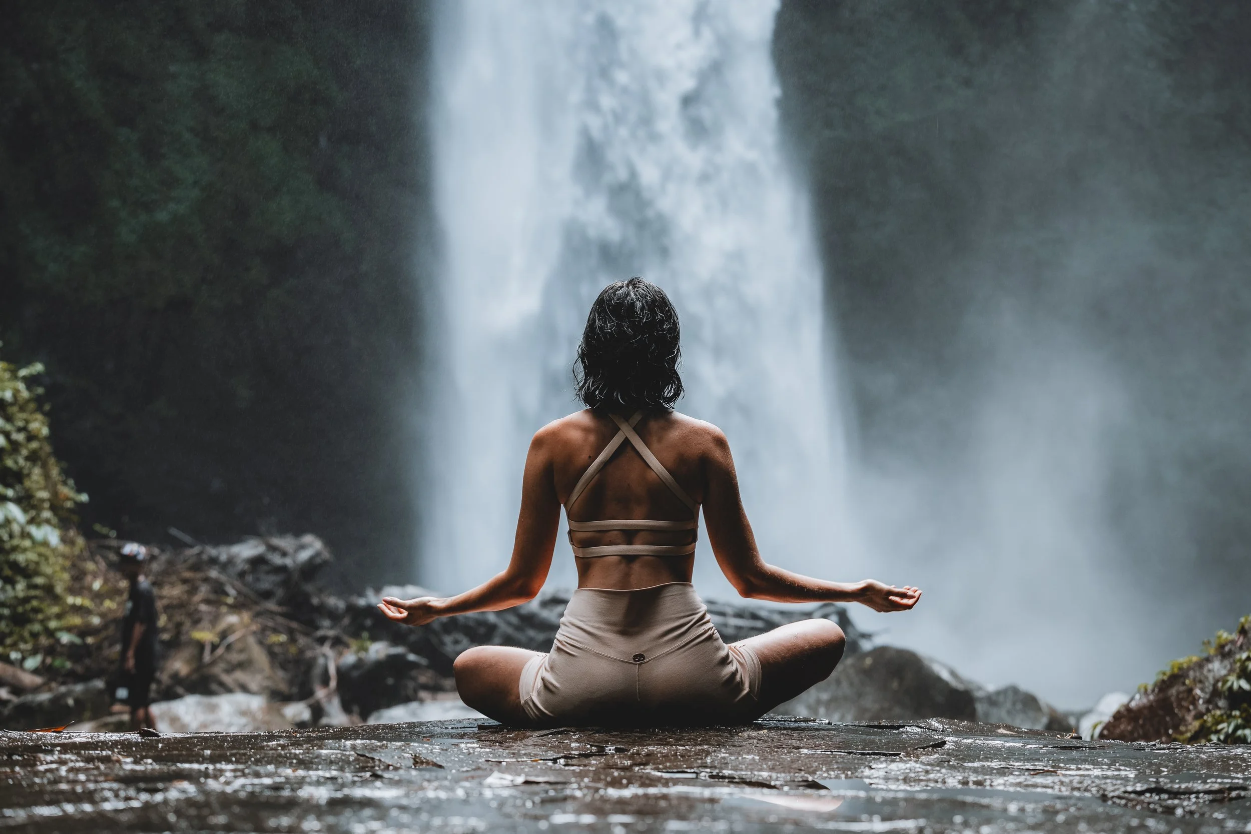 A woman practicing yoga in a seated position with crossed legs and arms extended, facing a waterfall surrounded by lush green foliage.