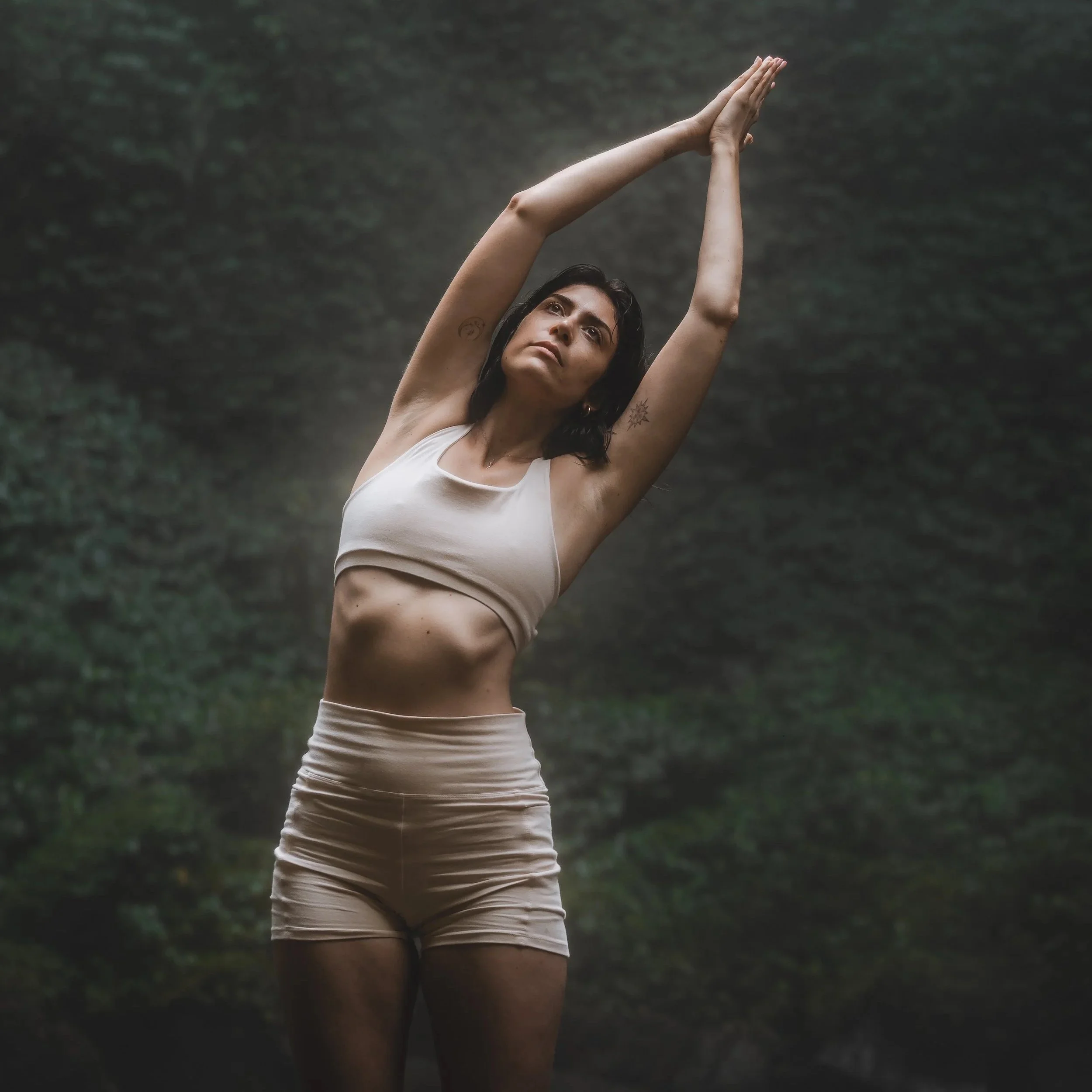A woman with dark hair and tattoos on her left arm practicing yoga outdoors, raising her arms above her head in a standing pose, against a blurred green background.