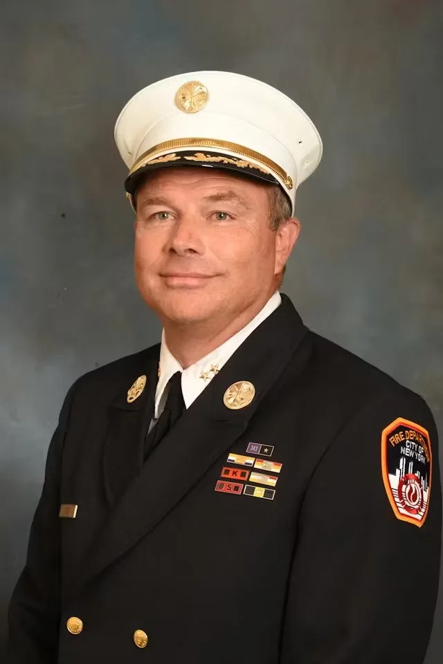 A fire department officer in a formal uniform standing in front of a brick wall and emblem.