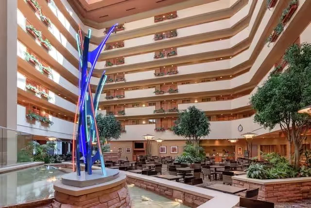 Atrium of a multi-story hotel with a colorful sculpture, dining area, indoor trees, and balconies.