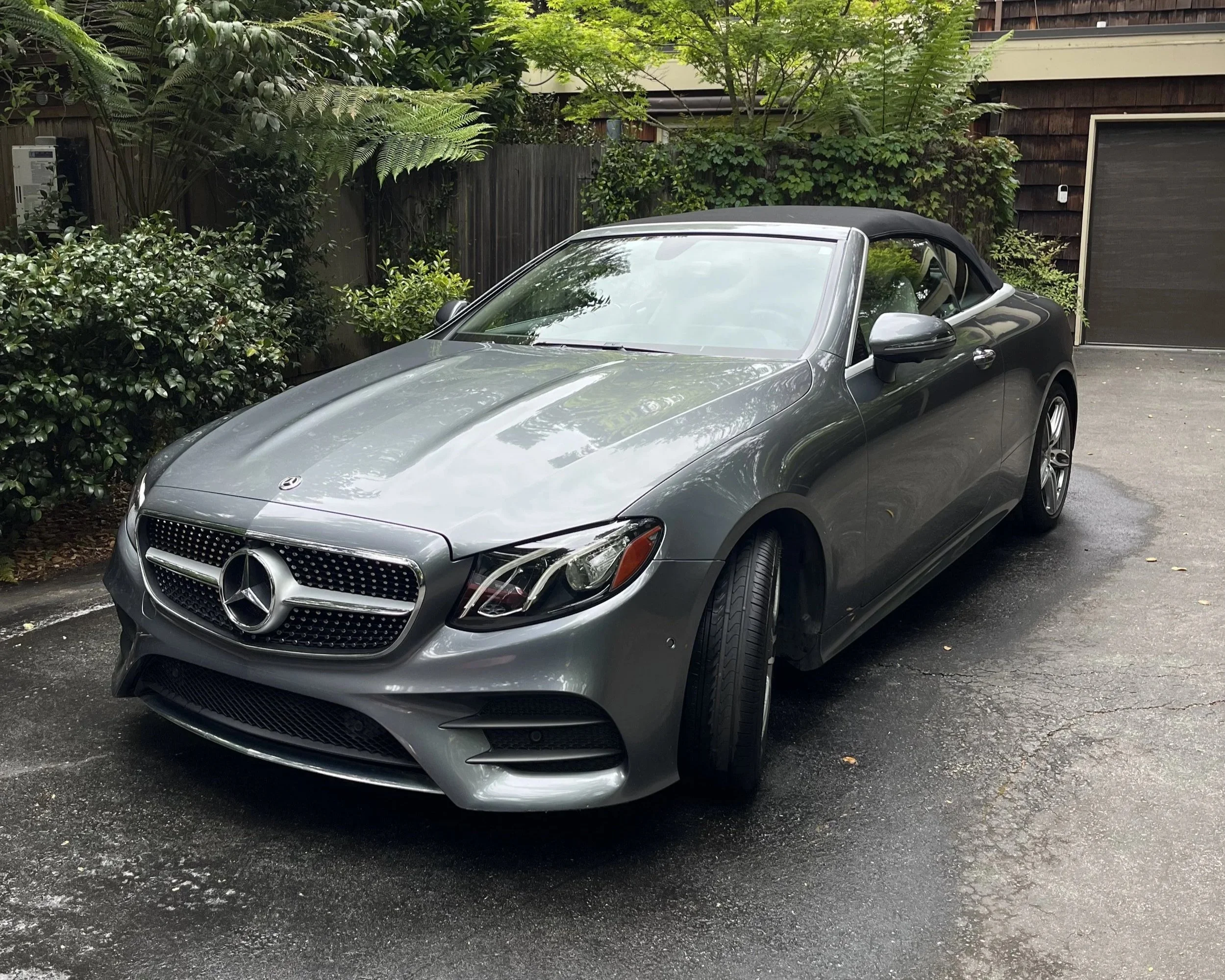 A gray Mercedes-Benz convertible car with a black soft top parked on a driveway surrounded by greenery.