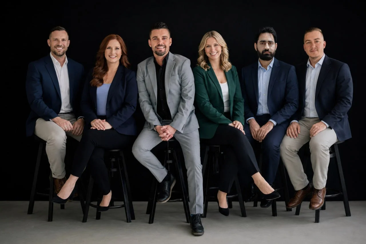 Group of six diverse professionals sitting on stools against a black background, dressed in business casual attire, smiling at the camera.