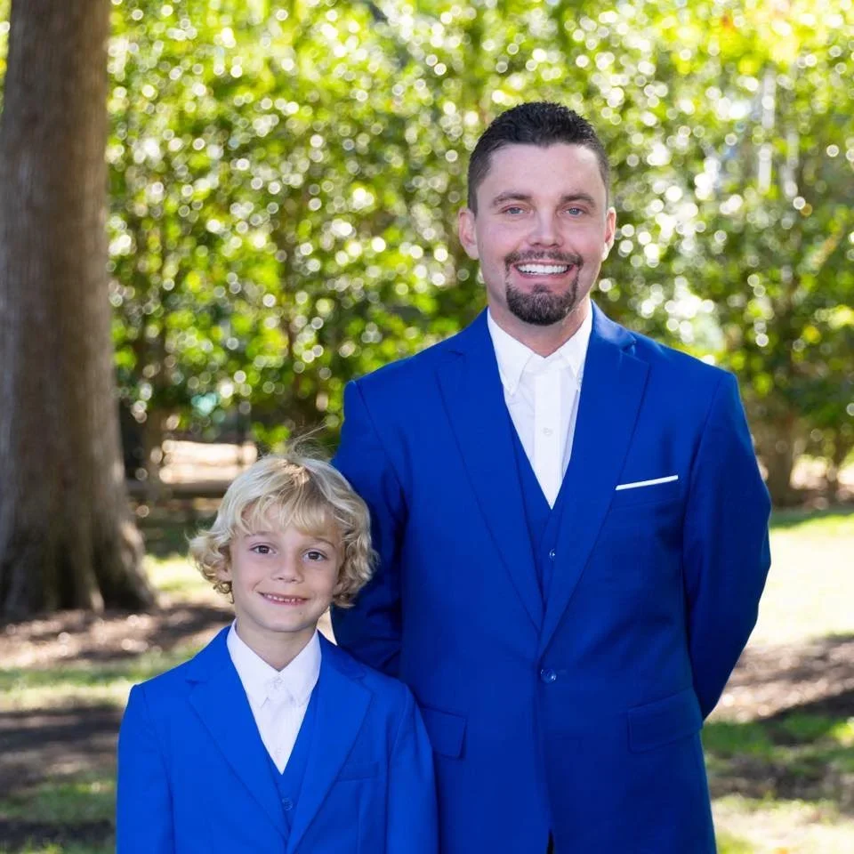 A man in a blue suit and a young boy in a matching blue suit pose outdoors in front of trees, smiling at the camera.